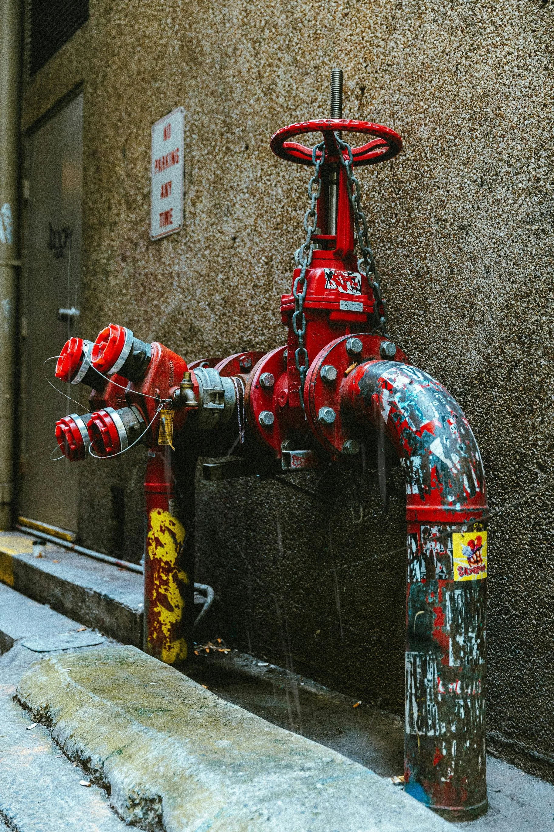 A red fire hydrant covered with colorful stickers, standing against a textured beige wall, with a yellow parking block in the foreground.
