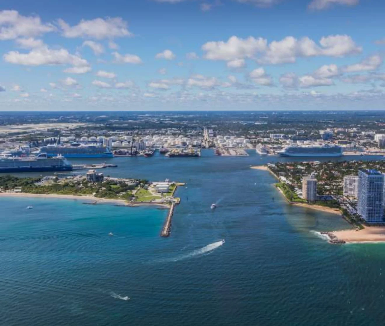 Aerial view of a coastal city with a large waterway, ships, high-rise buildings, and beaches under a partly cloudy sky.