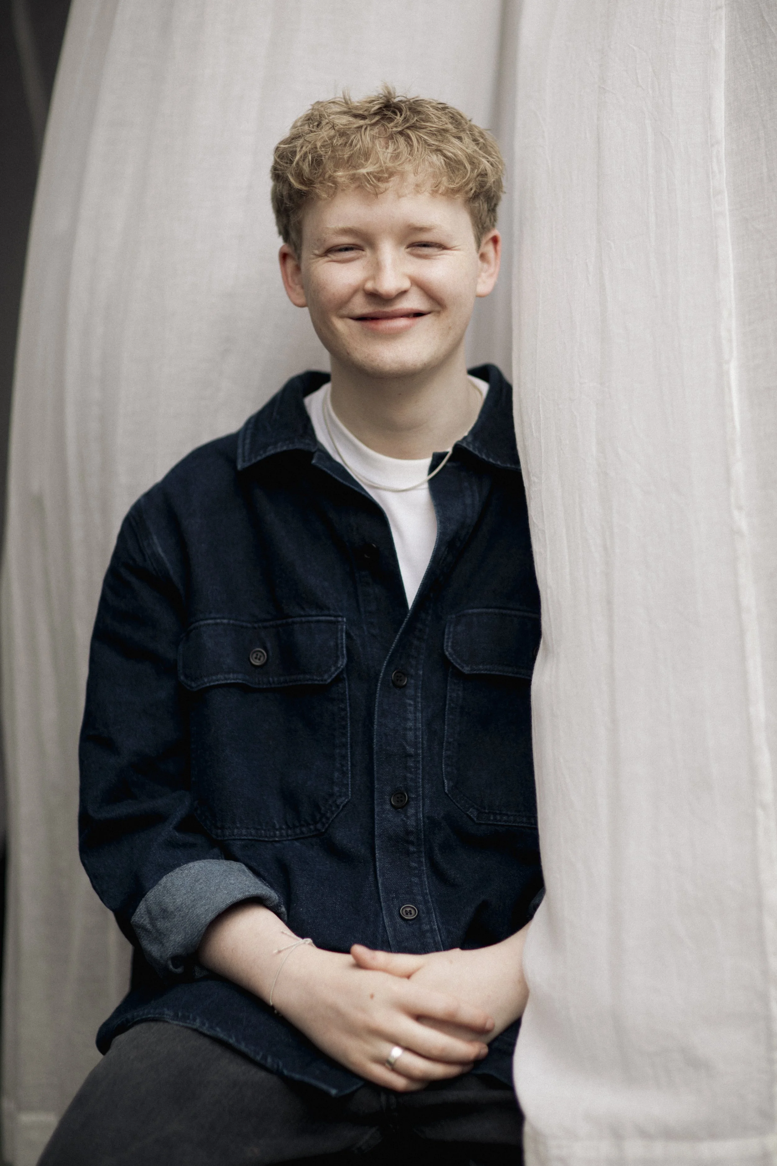 A smiling young man with curly blond hair, wearing a dark denim jacket over a white shirt, sitting with his hands clasped, partially obscured by a light-colored curtain.