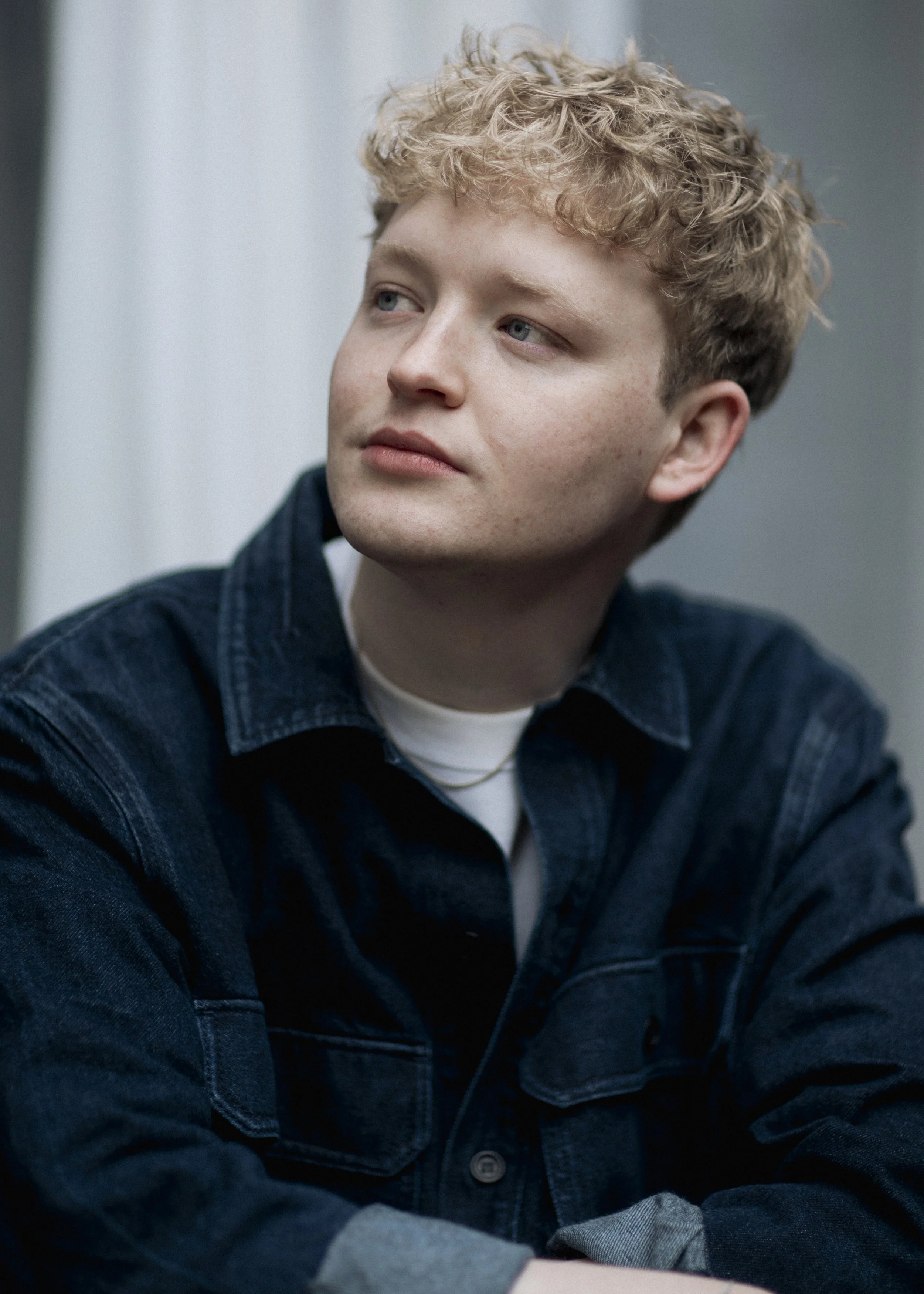 A young man with curly blond hair and blue eyes wearing a dark denim jacket and a white shirt looks to the left.