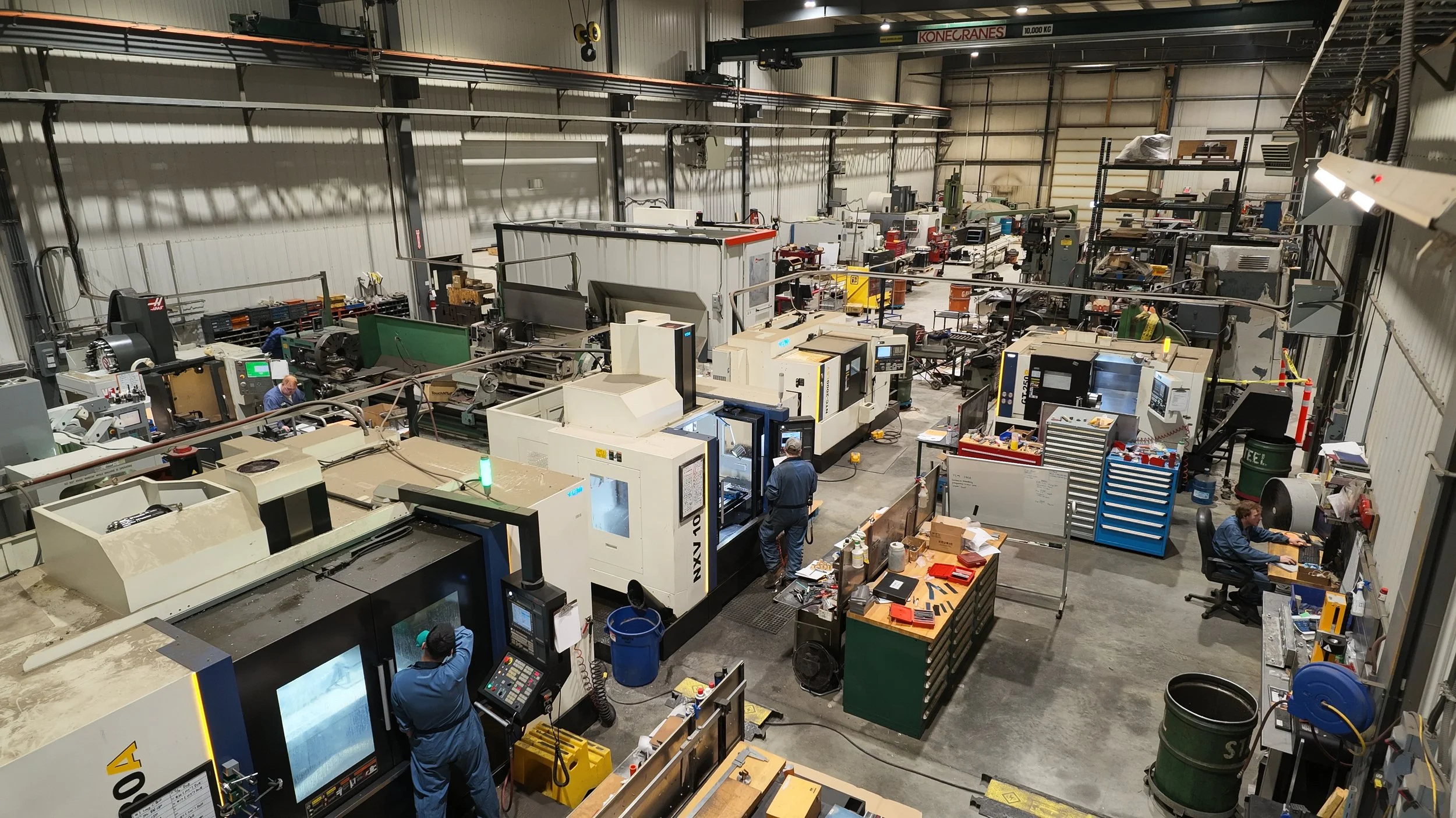 An industrial manufacturing facility with various machines, workstations, and workers engaged in tasks. Large equipment and tools are visible, along with storage shelves and scattered supplies.