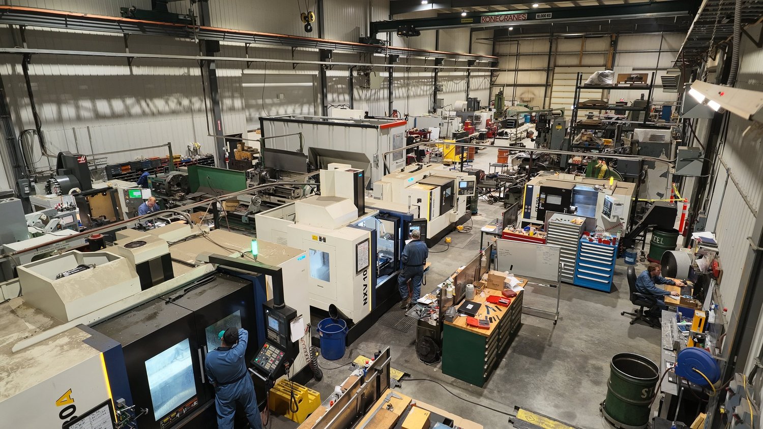 An industrial manufacturing facility with various machines, workstations, and workers engaged in tasks. Large equipment and tools are visible, along with storage shelves and scattered supplies.