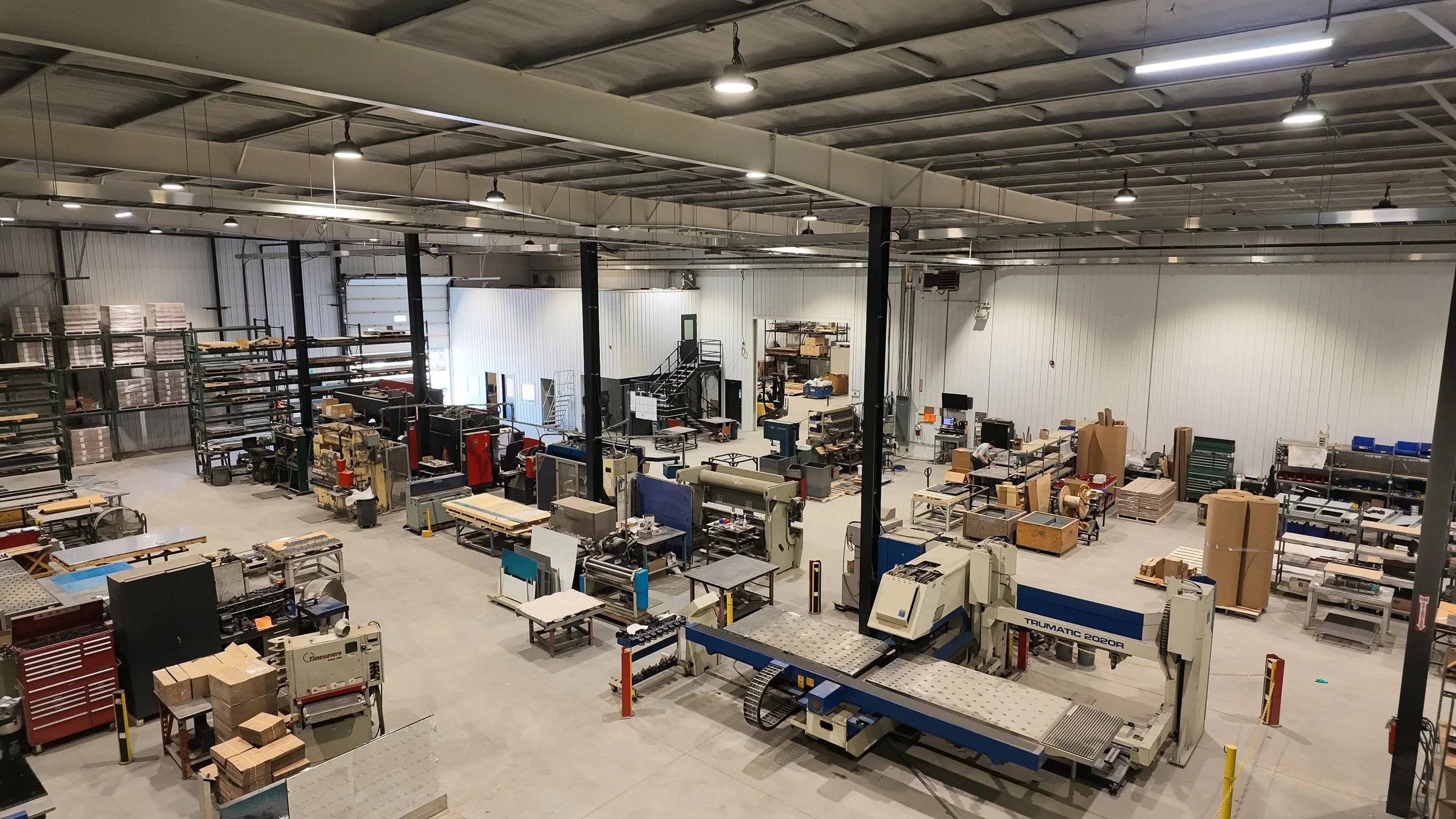 A large industrial warehouse filled with machinery, workbenches, and shelves with supplies, illuminated by ceiling lights.