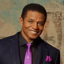 Portrait of a smiling man in a dark suit with a purple tie and pocket square, standing against a textured background.