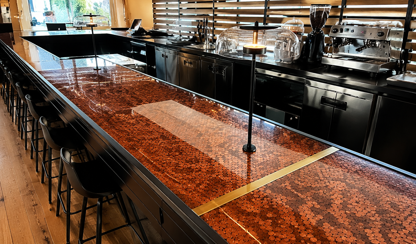 Modern bar or restaurant counter with mosaic tile surface and black seating, equipped with coffee machine and food display cases, in a well-lit setting.