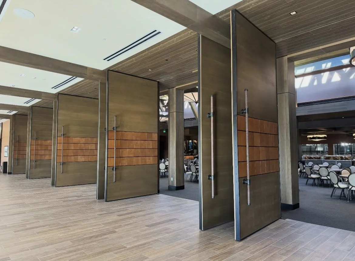 Large, modern restaurant or event space with wooden flooring, wooden walls, and big barn-style doors that function as entrances. Inside, there are tables and chairs for dining, with a mix of light and dark wood accents and natural light coming in from large windows.