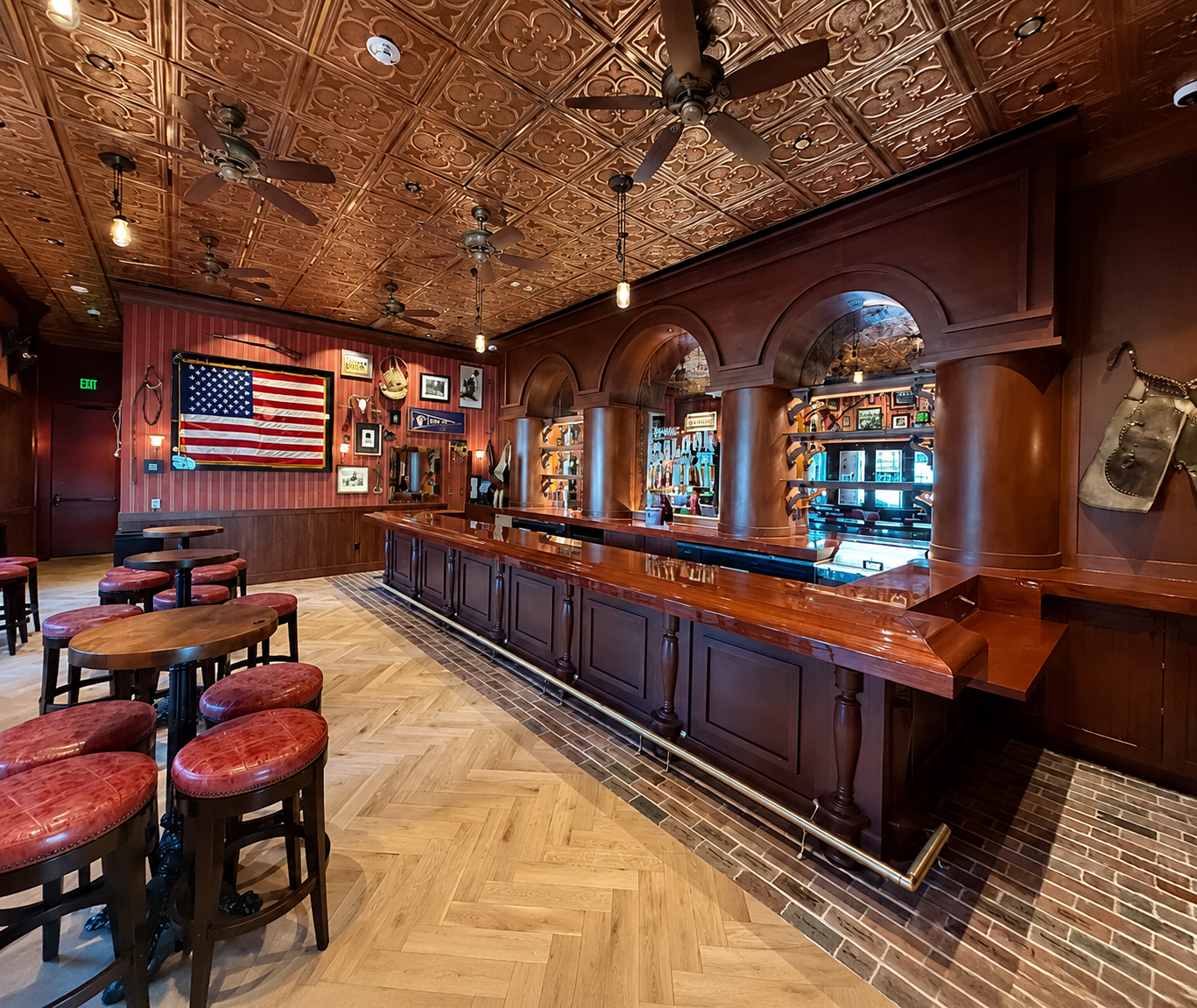 An empty bar with a wooden counter, bar stools with red cushions, and a wood-paneled area decorated with pictures, flags, and outdoor equipment. The ceiling has ornate wooden tiles and ceiling fans.