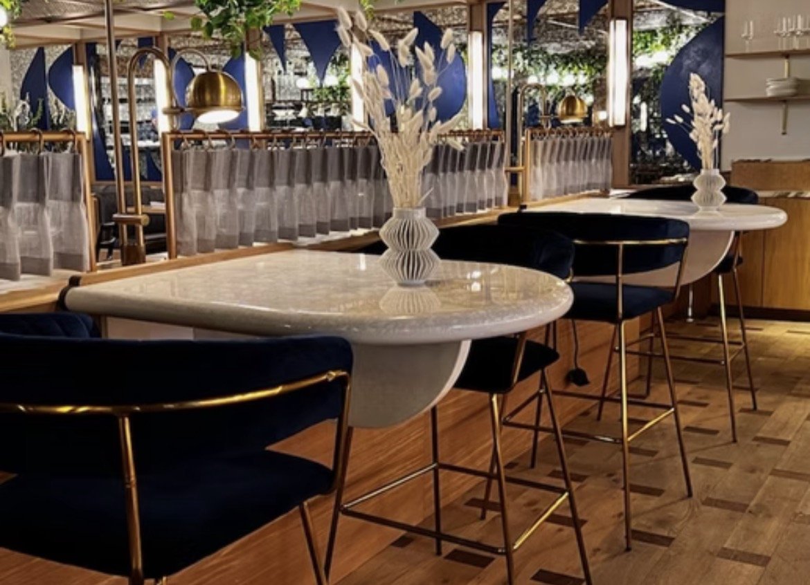 Interior of a modern restaurant with marble-topped tables, navy blue velvet chairs with brass frames, white vases with white decorative flowers, and a wooden floor. The background features a wooden partition with serving areas, blue wall art, and gre