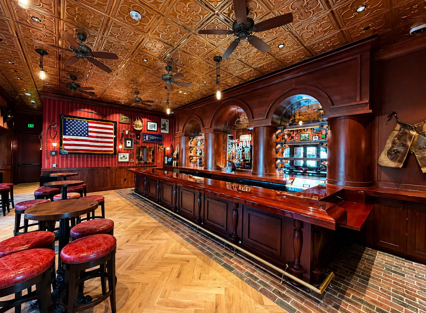 Interior of a bar with a polished wooden counter, red upholstered barstools, wooden tables and chairs, a red striped wall decorated with an American flag, framed pictures, and vintage items, with a decorative wooden ceiling and ceiling fans.
