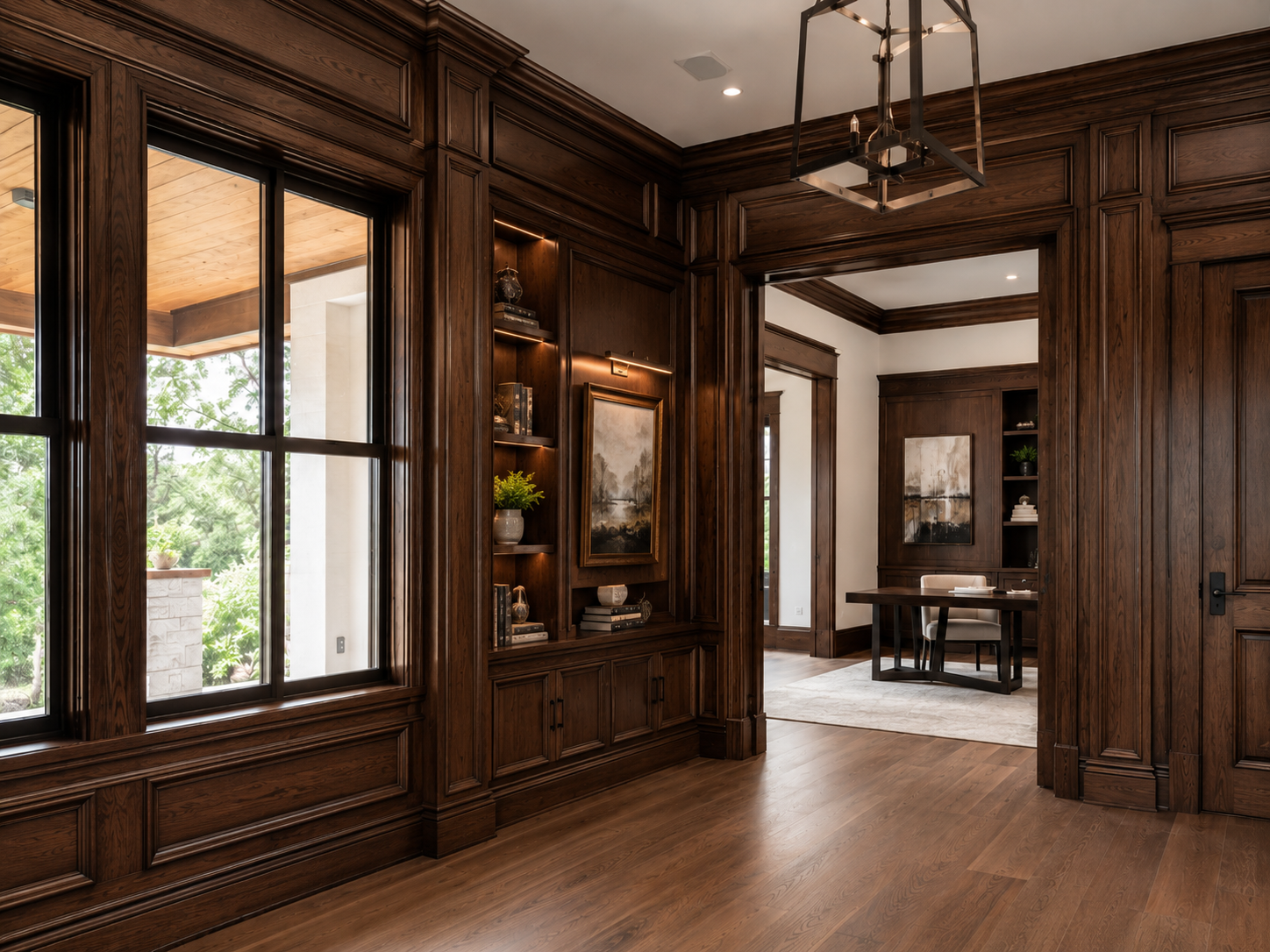 A room with dark wooden paneling and trim, large window with divided panes showing greenery outside, built-in shelves with books, a potted plant, and decorative items, a painting on the wall, and doorways leading to other rooms with similar dark wood accents and a desk with chairs.