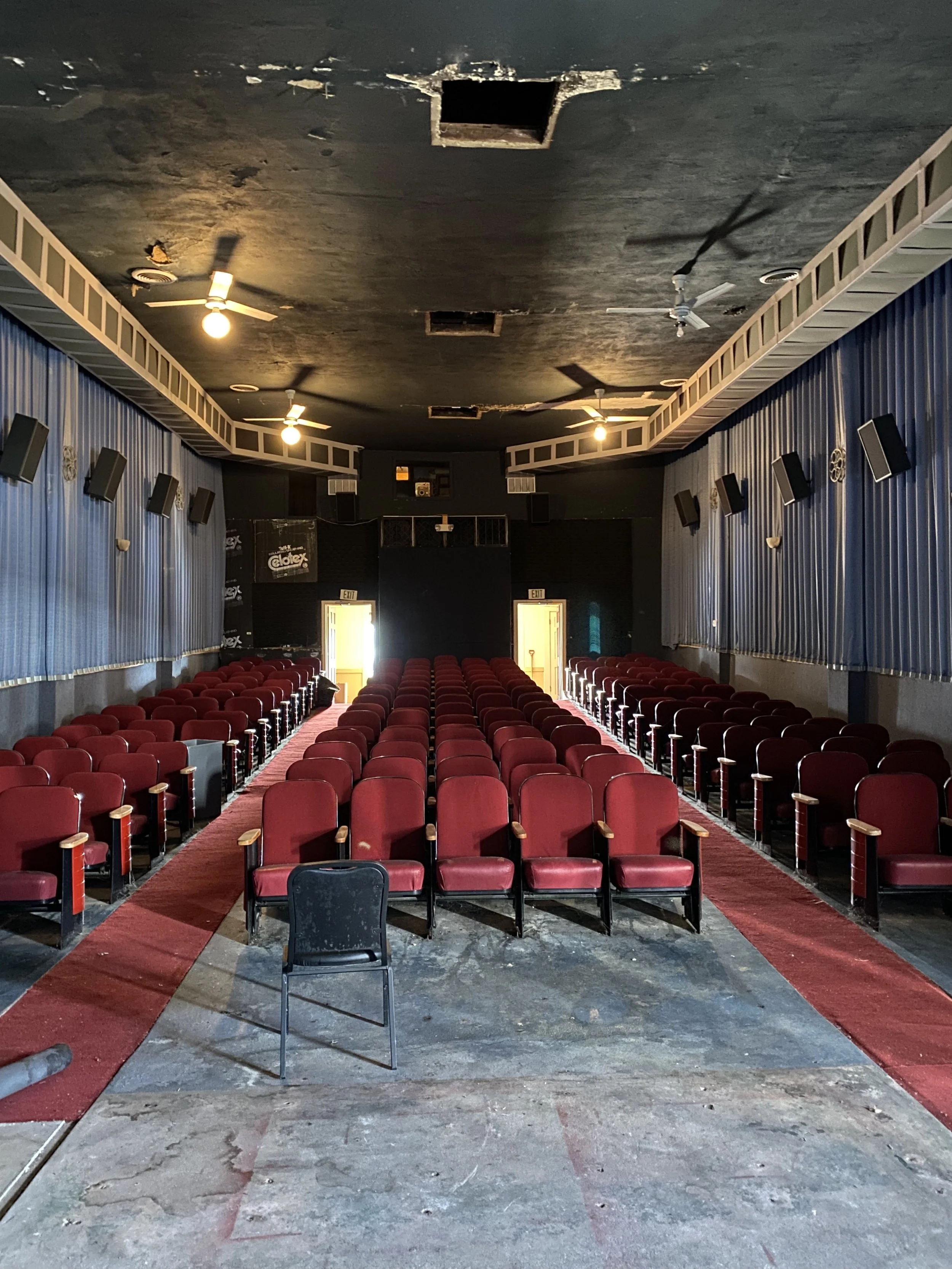 Empty auditorium with red chairs, a black chair in the front, blue curtains on the sides, ceiling fans, and a damaged floor.
