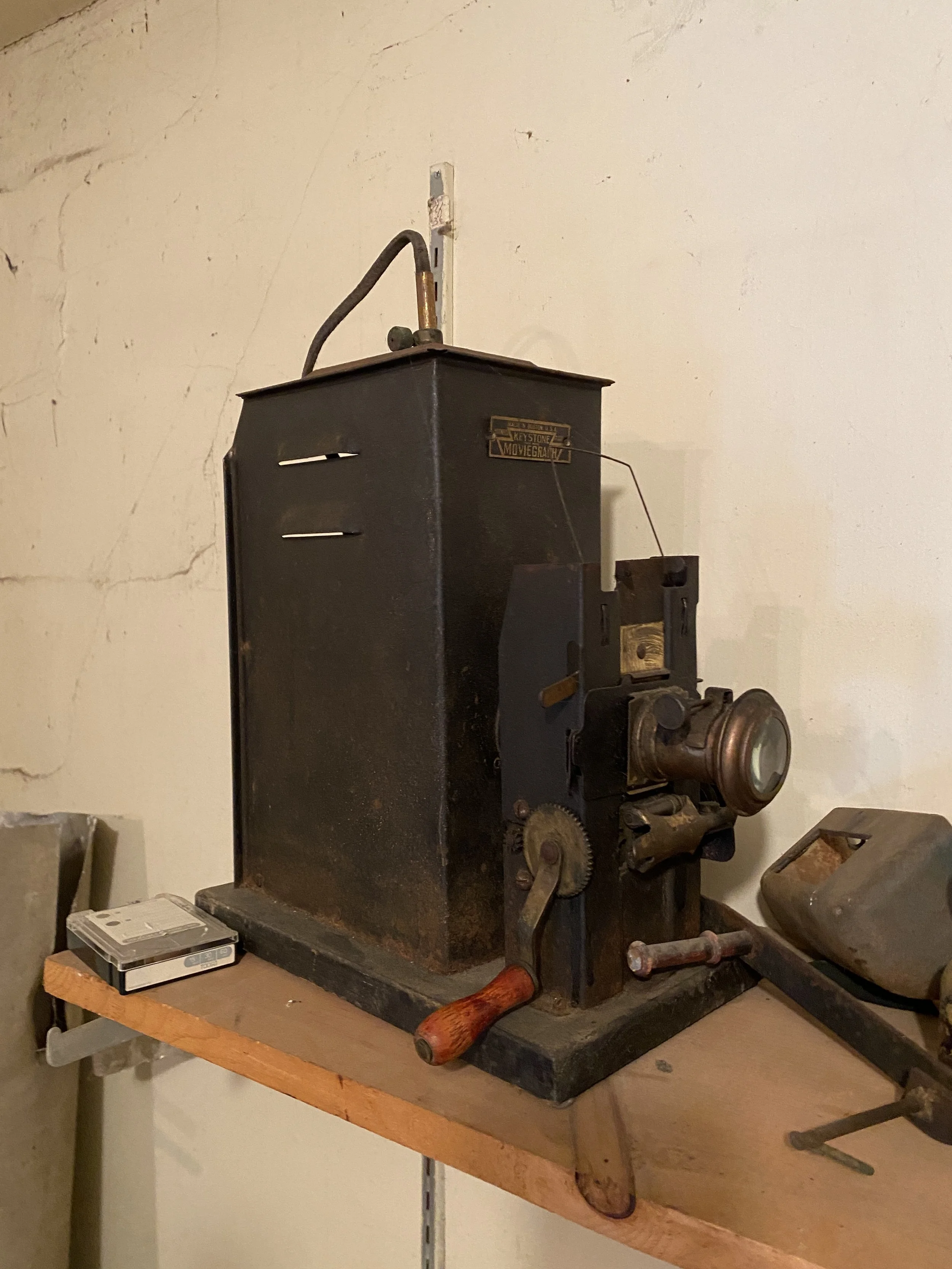 An antique film projector on a wooden shelf, with a cast iron body, gears, a handle, and a lens, placed against a plaster wall.