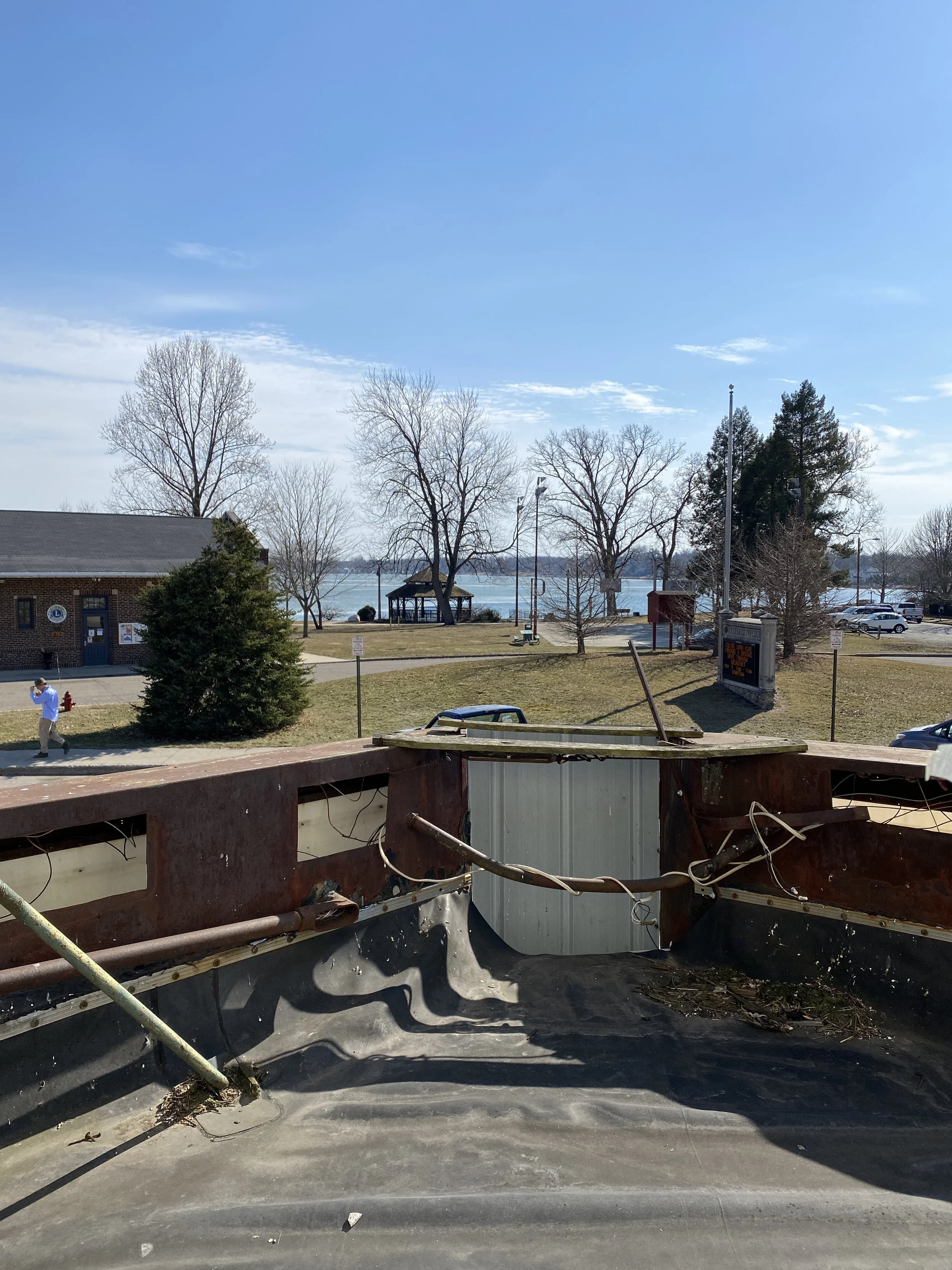 View from inside a rusted, wrecked vehicle facing a park with trees, a gazebo, and a lake in the background on a clear day.