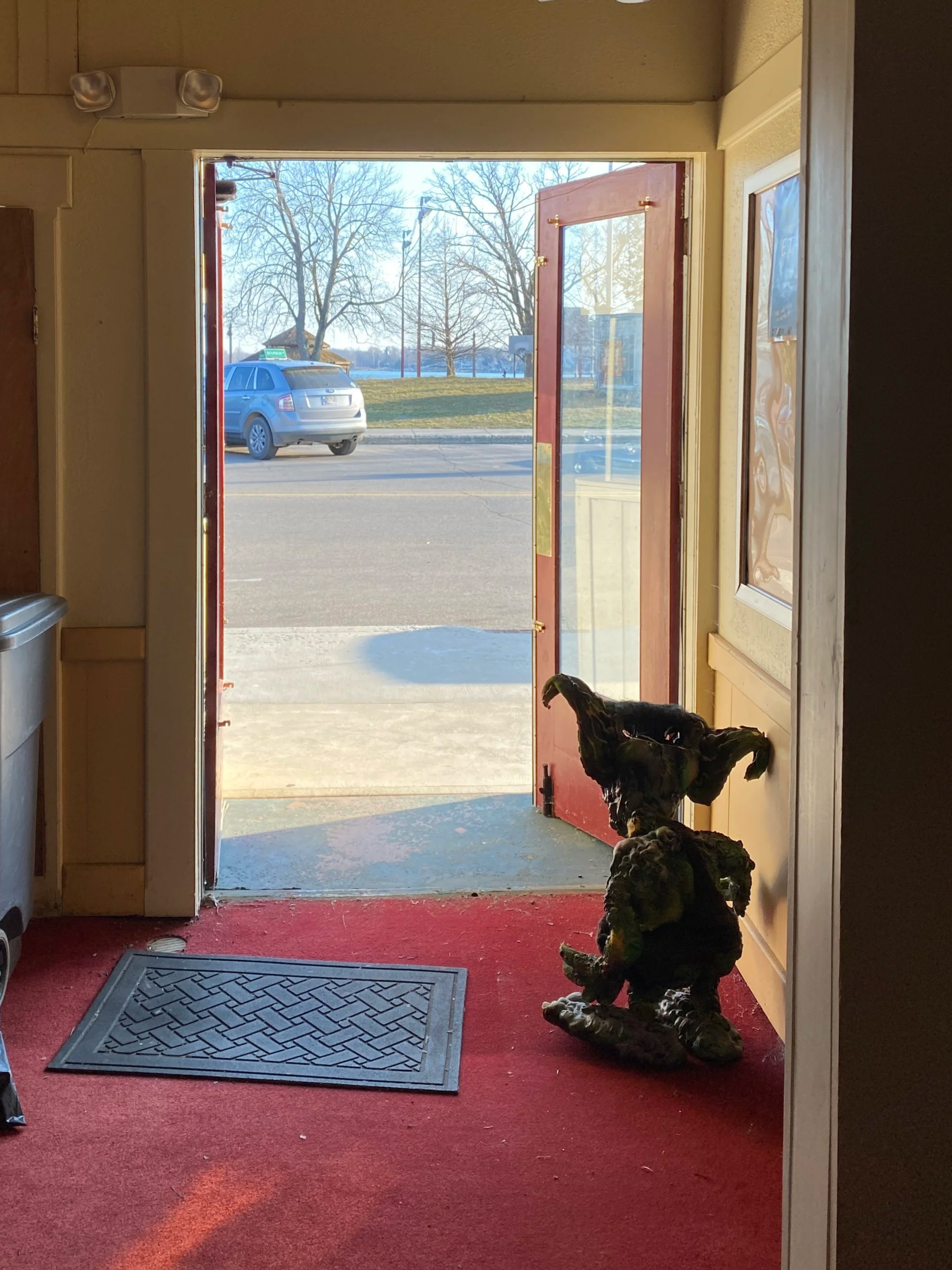 Inside view of a building with a red carpet entrance, a black statue of a dog sitting on the floor, a rubber doormat, and a view of the street outside with a parked blue car, leafless trees, and a field in the background.