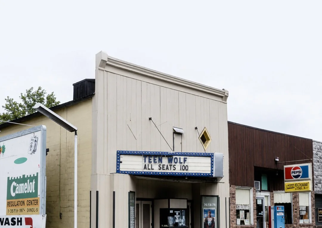 A small movie theater with a marquee that reads 'Teen Wolf All Seats 100' in a small town, surrounded by other storefronts and signs, including a Pepsi sign and a Camelot regulation center sign.