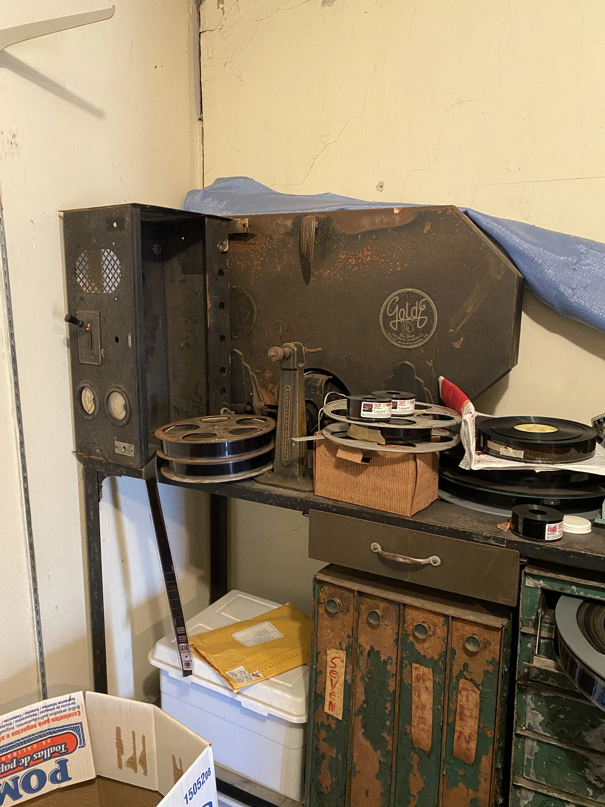 A cluttered workspace with an old, rusty mechanical device labeled 'Gold'. There are film reels, a roll of tape, and various boxes on the table, with a cardboard box in the foreground and a worn, green and orange metal cabinet underneath.