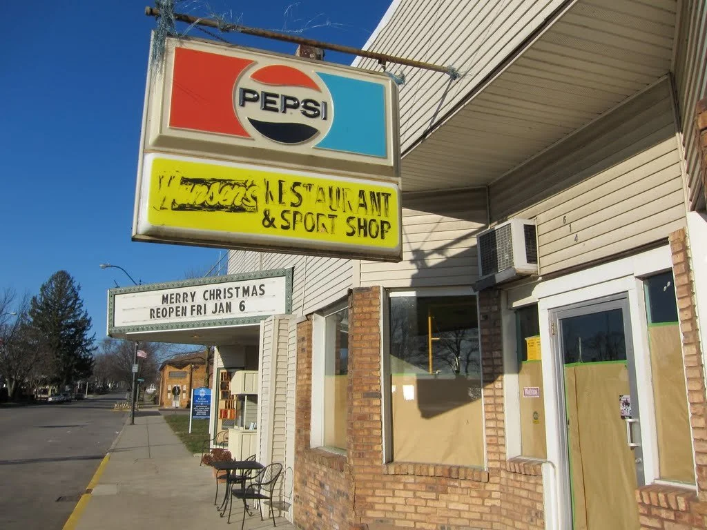 A street view of a small business with signs reading Pepsi, restaurant & sport shop, and a message about reopening after Christmas on a marquee. The building has brick and siding exterior, with a window covered by brown paper, and a door with a small window also covered. An outdoor chair is positioned near the entrance, and the street is lined with a sidewalk and some trees in the distance under a clear blue sky.