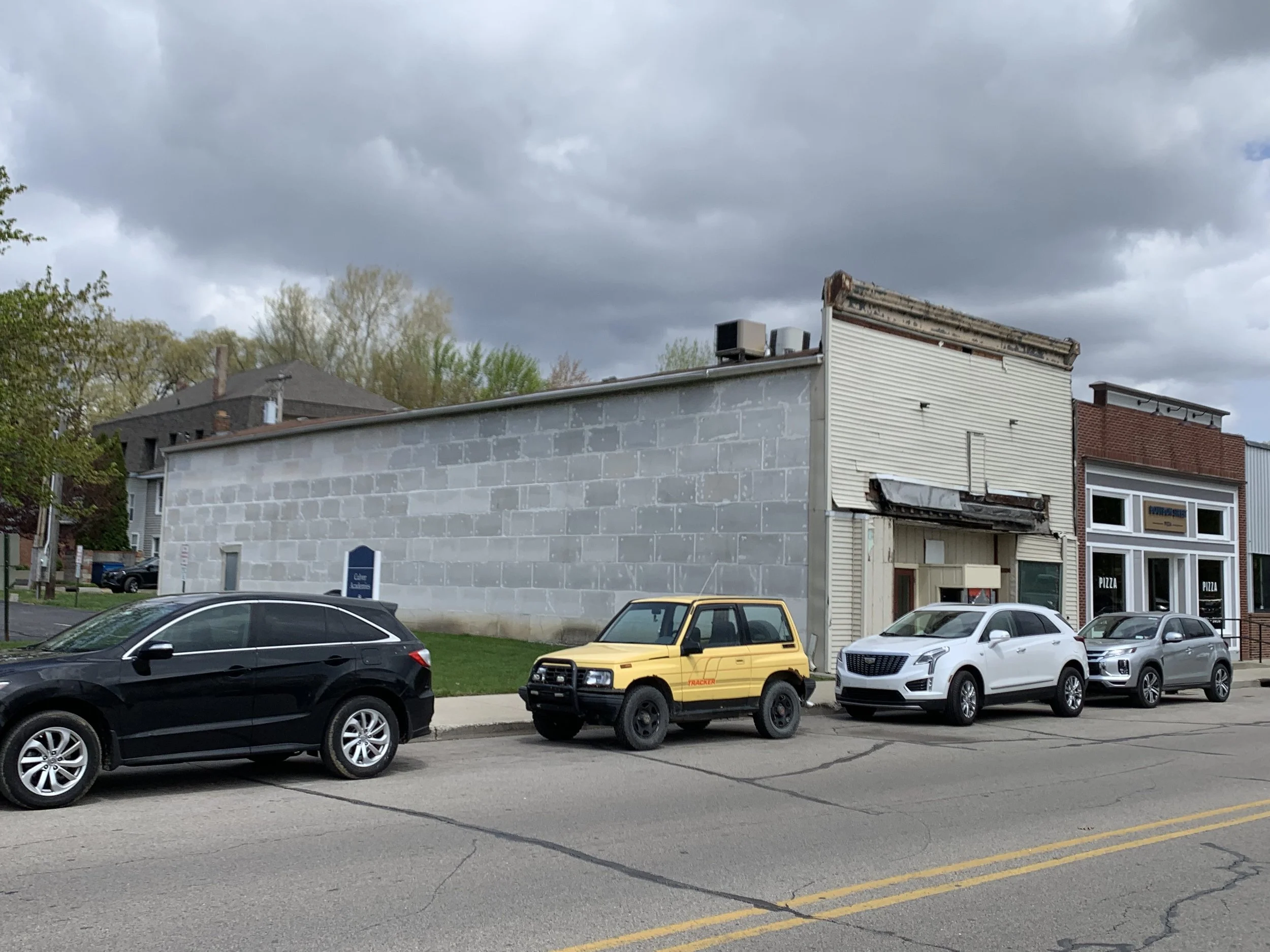 City street scene with parked cars in front of buildings under a cloudy sky.