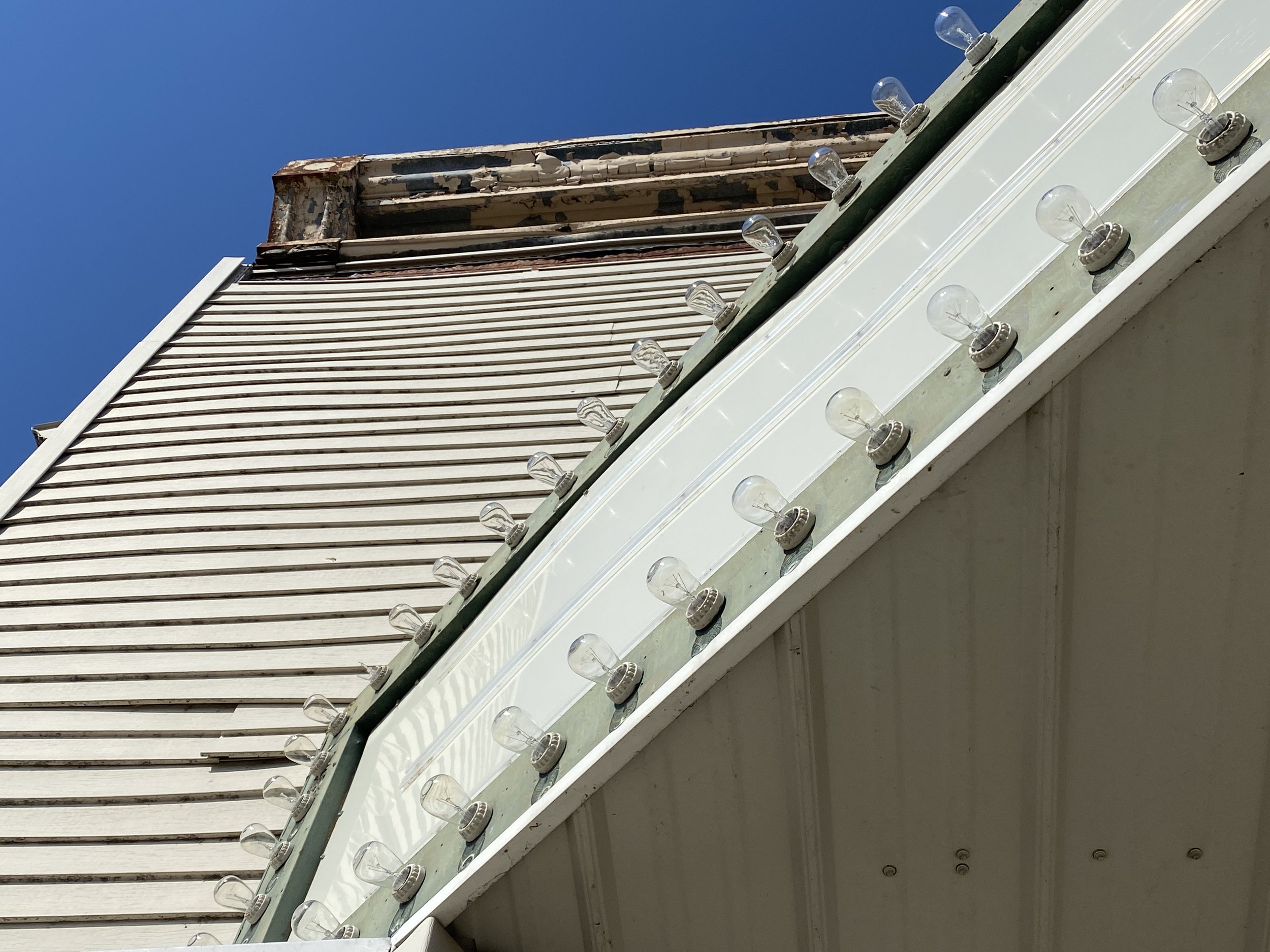 Close-up of old restaurant or motel sign with empty light bulbs, next to a building with horizontal white siding, under a clear blue sky.