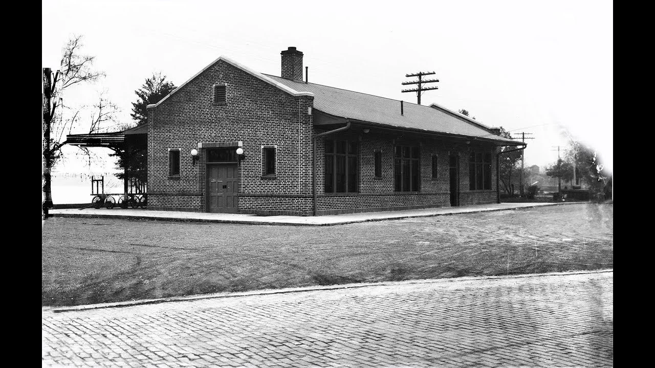 Black and white photo of a brick building with large windows, a chimney, and a covered porch, next to a cobblestone sidewalk and an unpaved area.