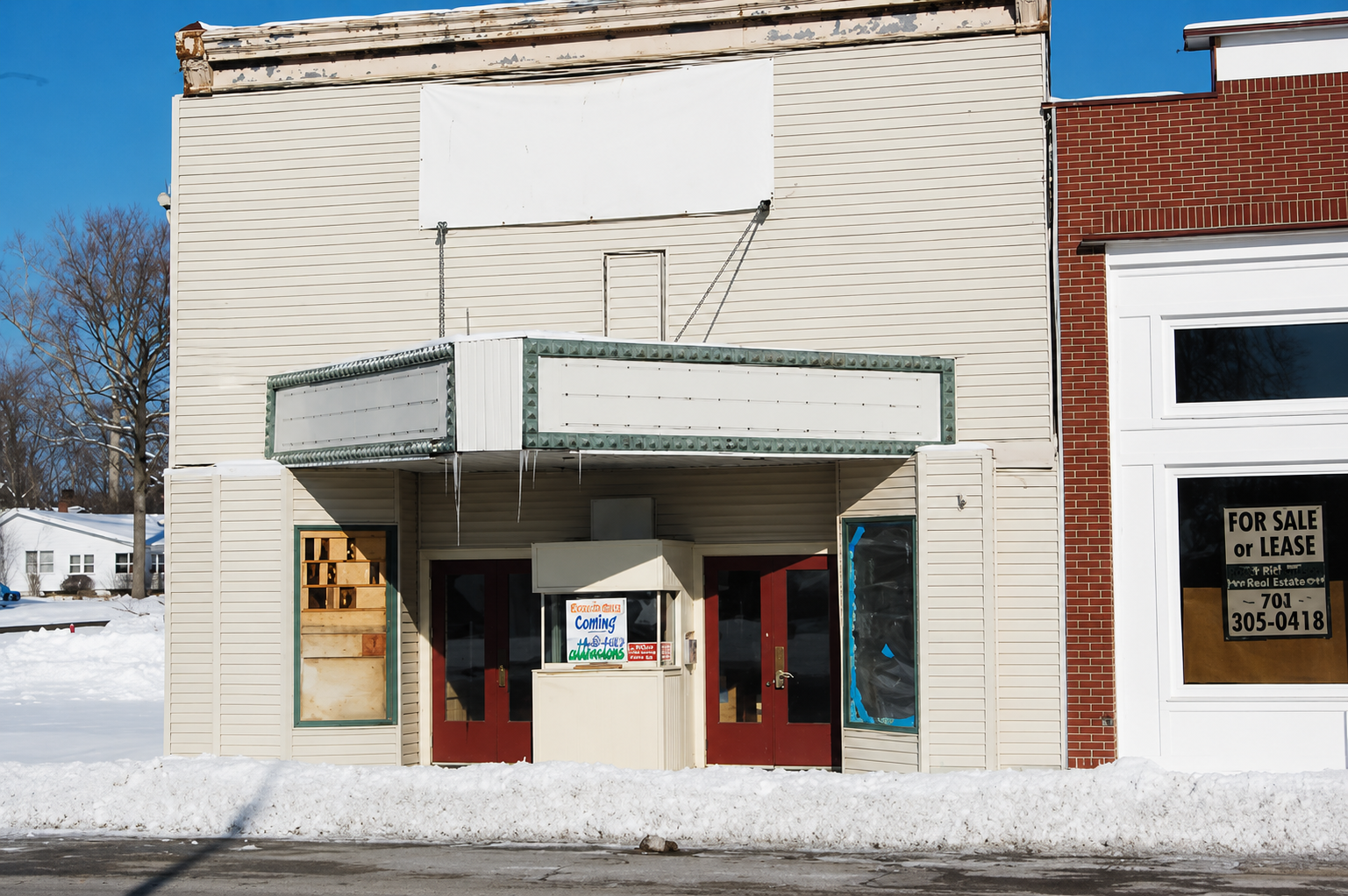Empty storefront with a marquee sign, boarded-up window, and a 'Coming Attractions' sign on the door. Snow on the ground and a neighboring red brick building with a 'For Sale or Lease' sign in the window.
