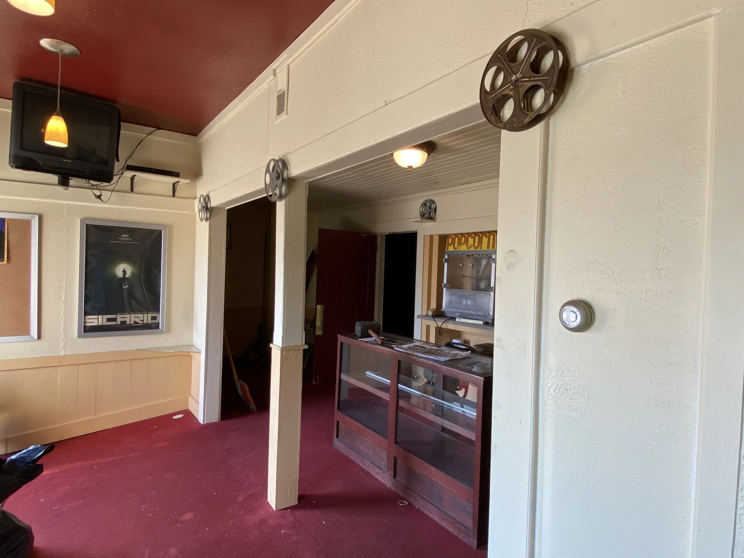 Interior view of a restaurant or movie theater lobby with a television on the wall, movie posters, a popcorn machine, display shelves, and decorative film reels hanging from the ceiling.