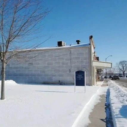 A snow-covered sidewalk next to a gray brick building with a blue sign, trees, and parked cars in the background on a clear winter day.