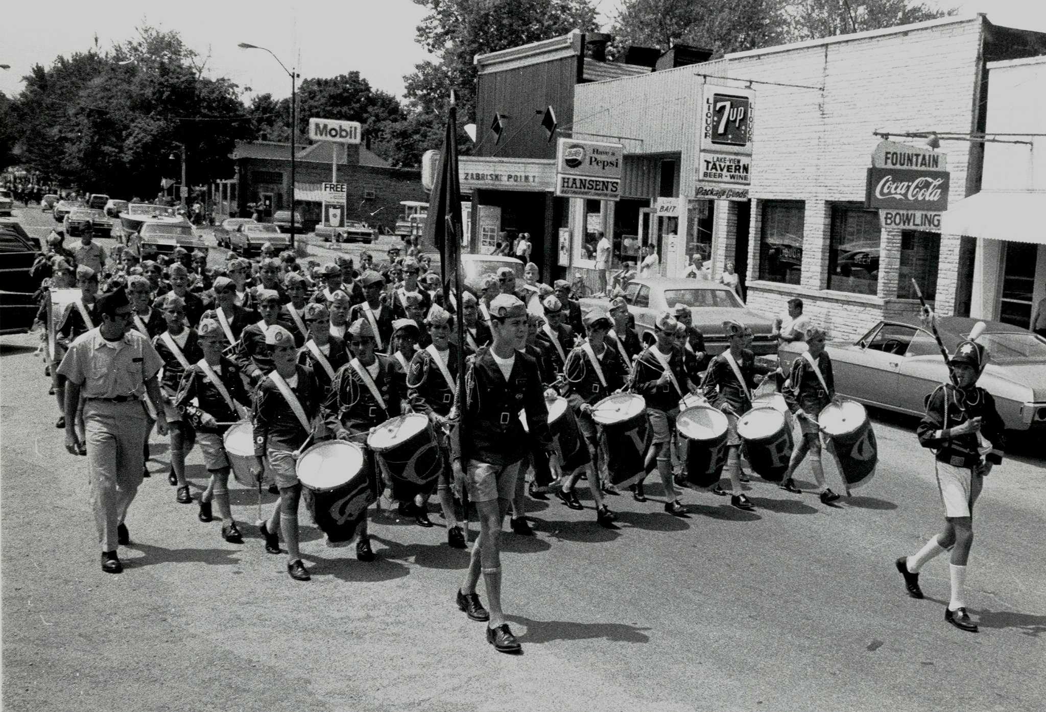 Black and white photo of a marching band with drum players leading a parade on a city street.