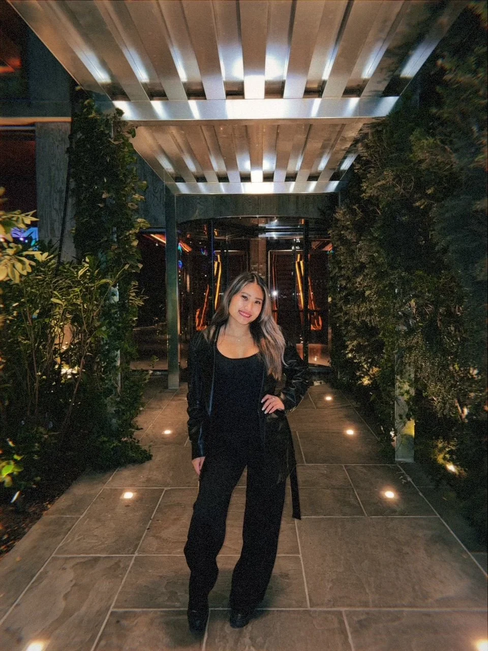 A woman wearing a black outfit and black coat, posing on a well-lit outdoor walkway at night, surrounded by green bushes and modern architectural elements.