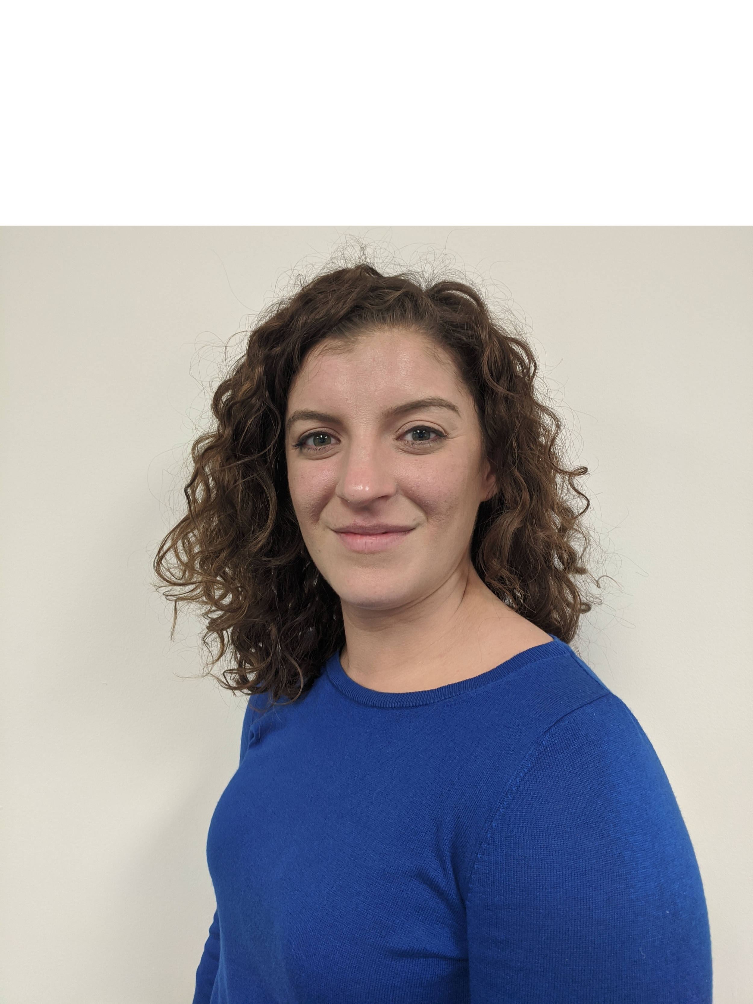 A woman with curly brown hair and blue eyes wearing a blue top standing against a plain white wall
