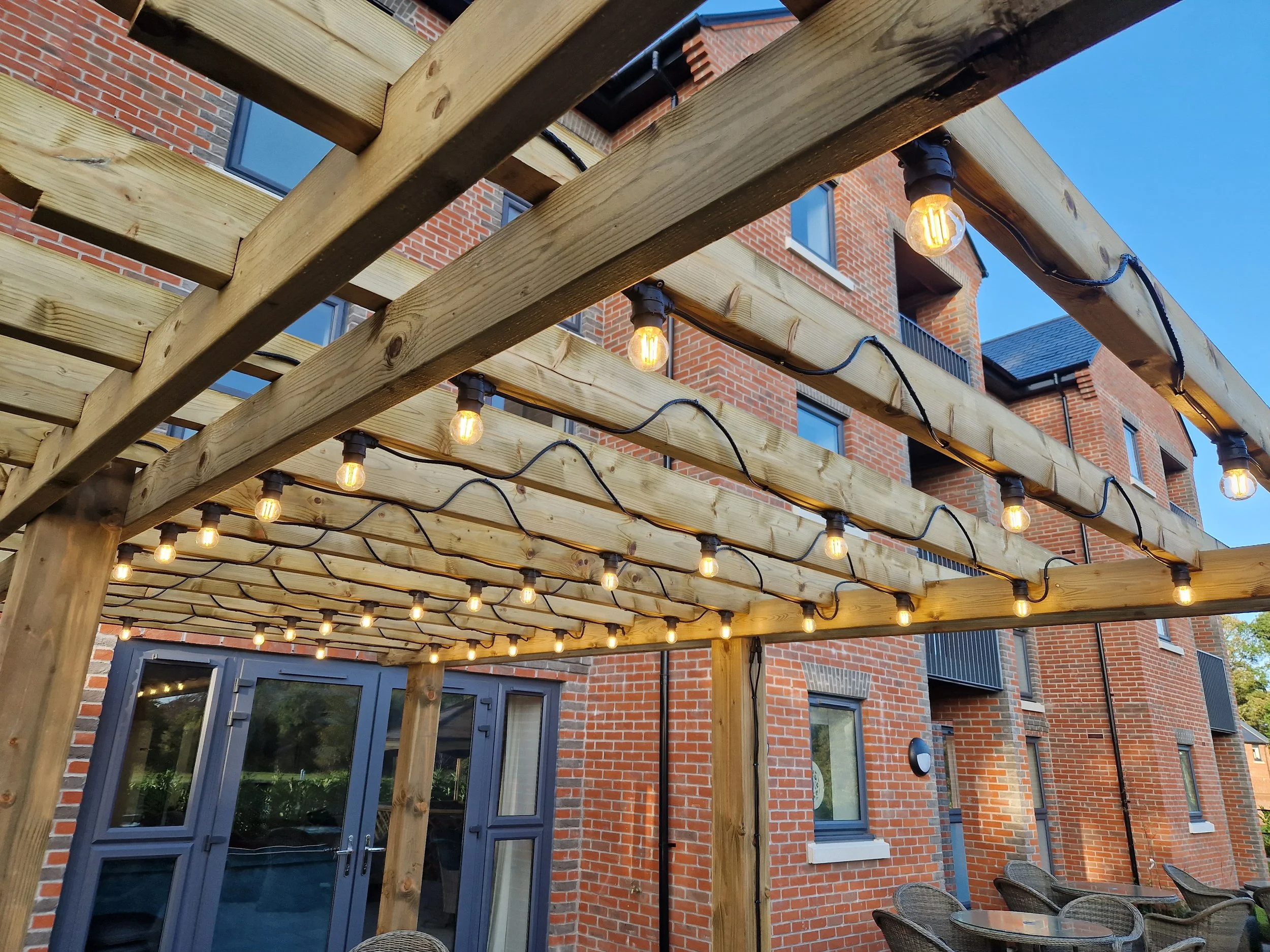 Wooden pergola with string lights over an outdoor seating area in front of a brick apartment building.