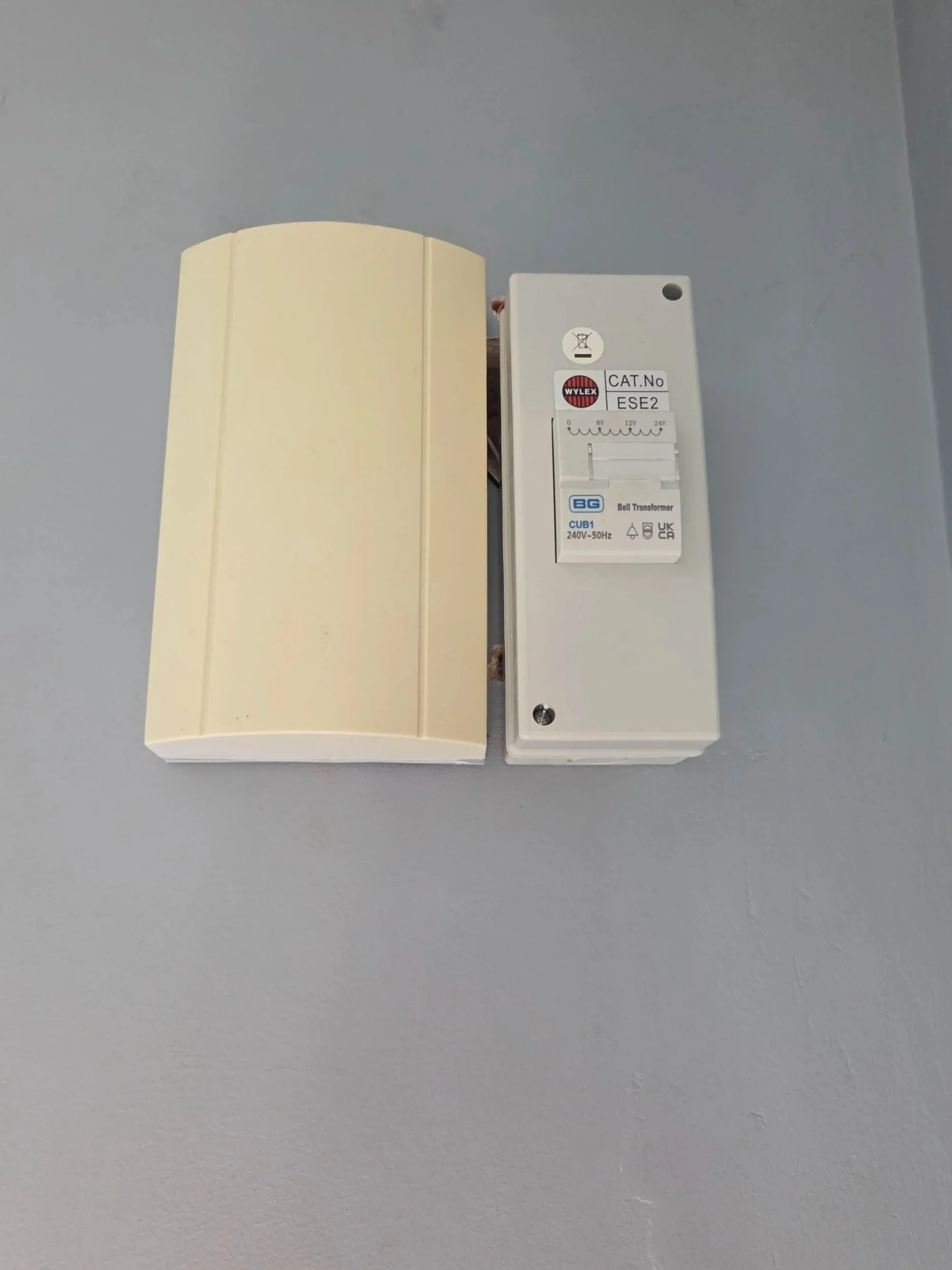 Close-up of an electrical transformer mounted on a wall, with a light-colored plastic protective cover beside it.