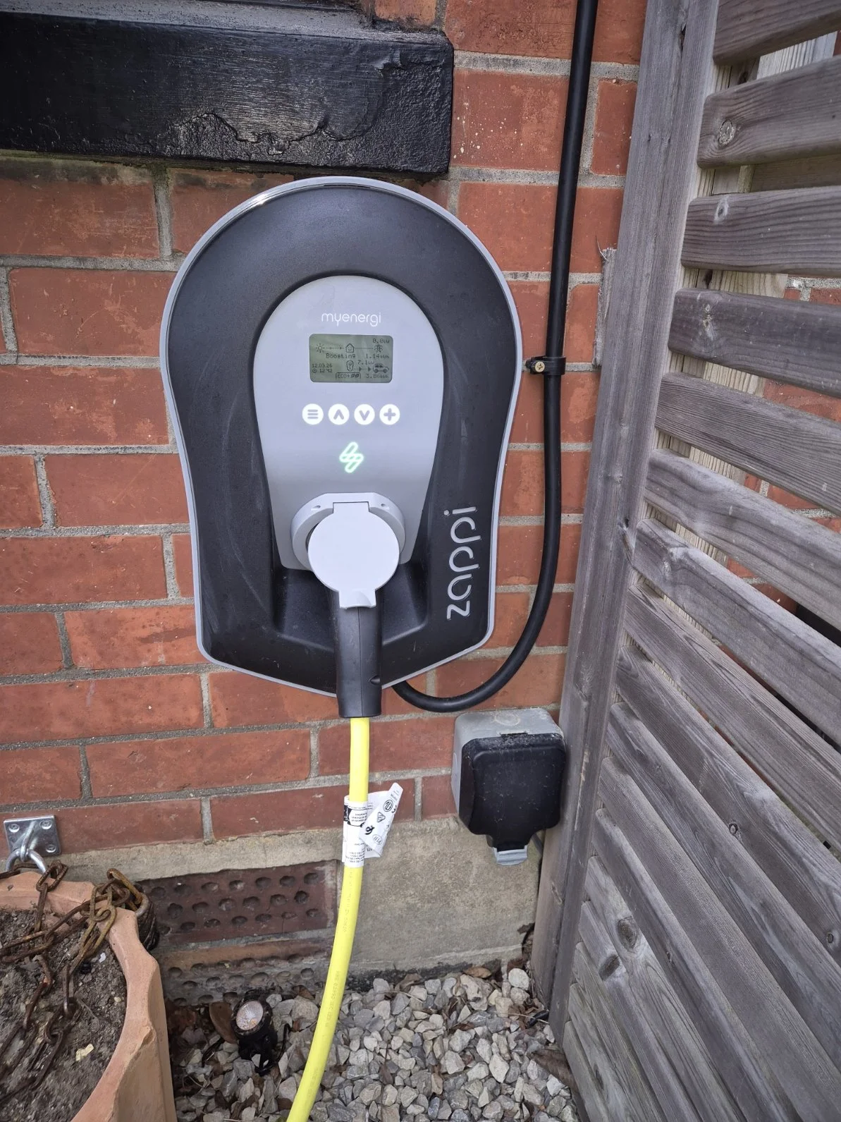 An electric vehicle charging station mounted on a brick wall, connected to a yellow charging cable, with a digital display and control buttons.