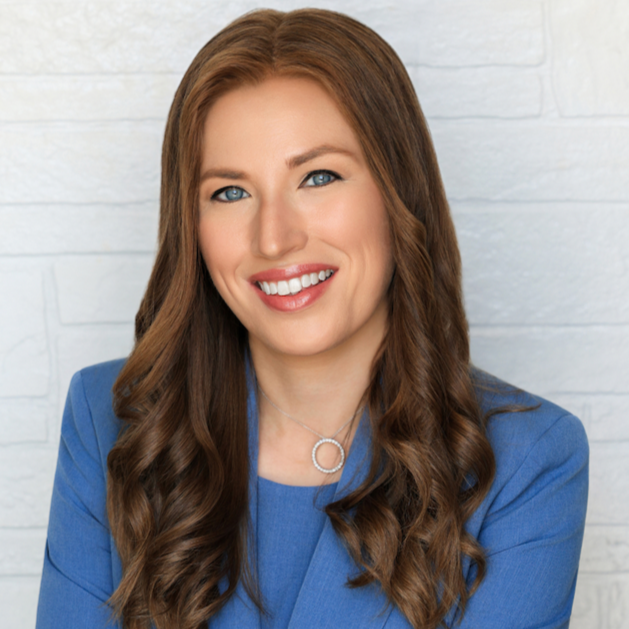 Headshot of a woman with long, wavy brown hair in a blue blazer, smiling against a white brick wall background.