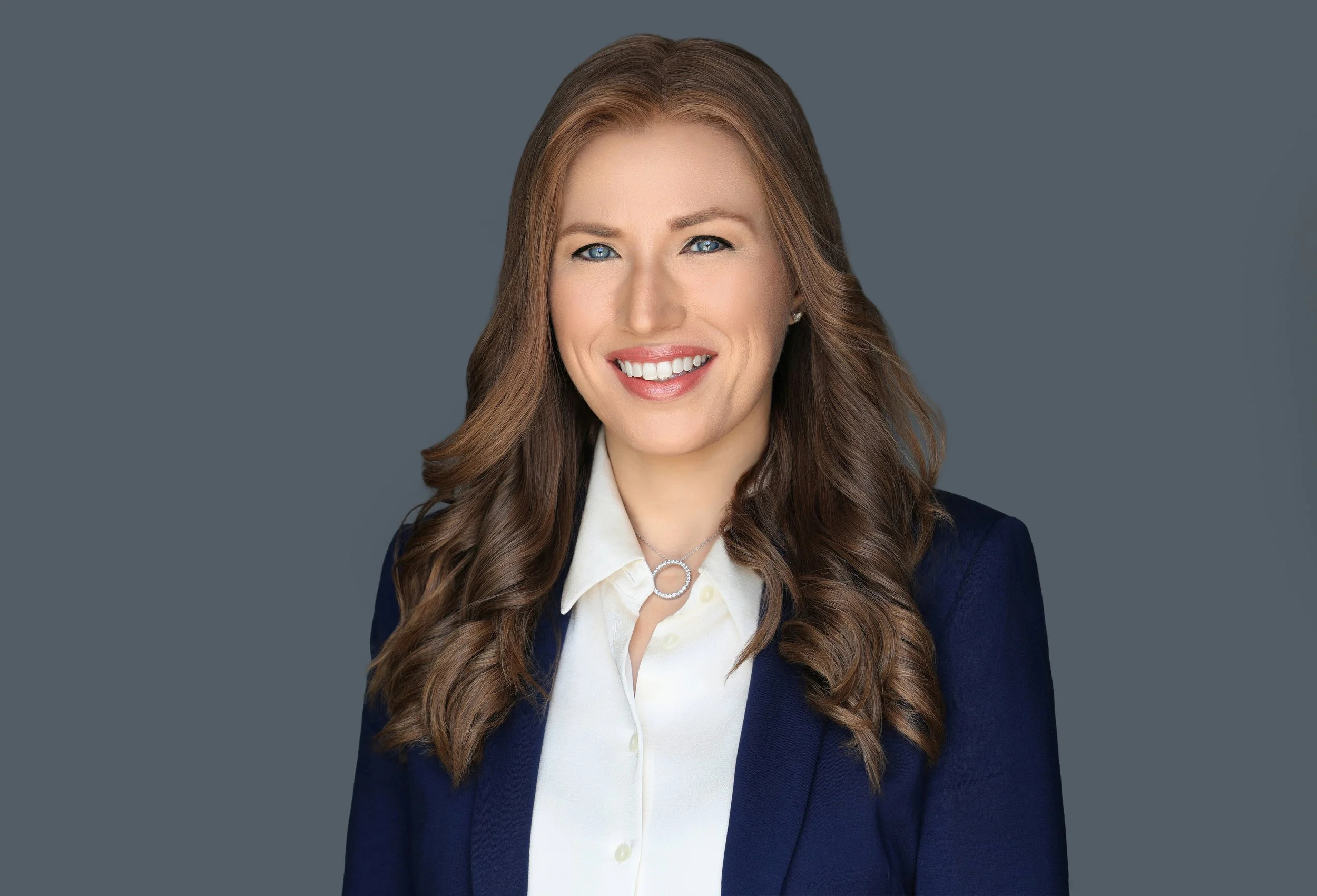 A woman with long wavy brown hair, blue eyes, and a bright smile, wearing a navy blazer and a cream blouse, standing against a gray background.