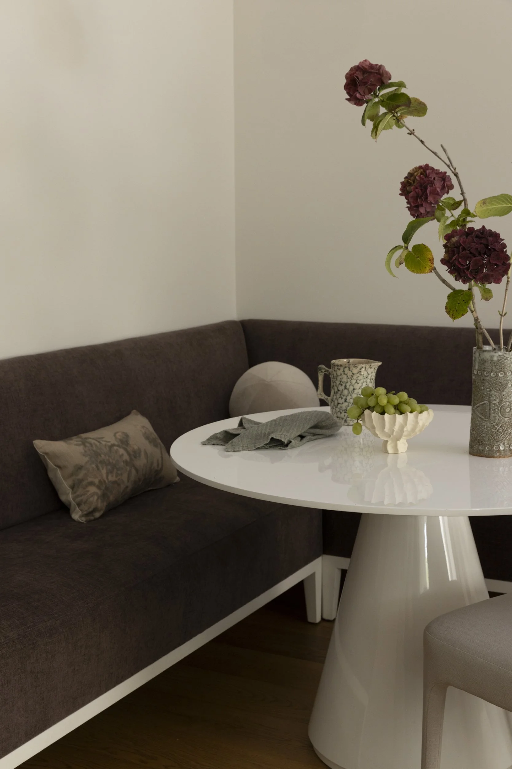 A round white table with a bottle of flowers, a bowl of grapes, and a mug, next to a brown sofa with a decorative pillow, in a room with plain walls.