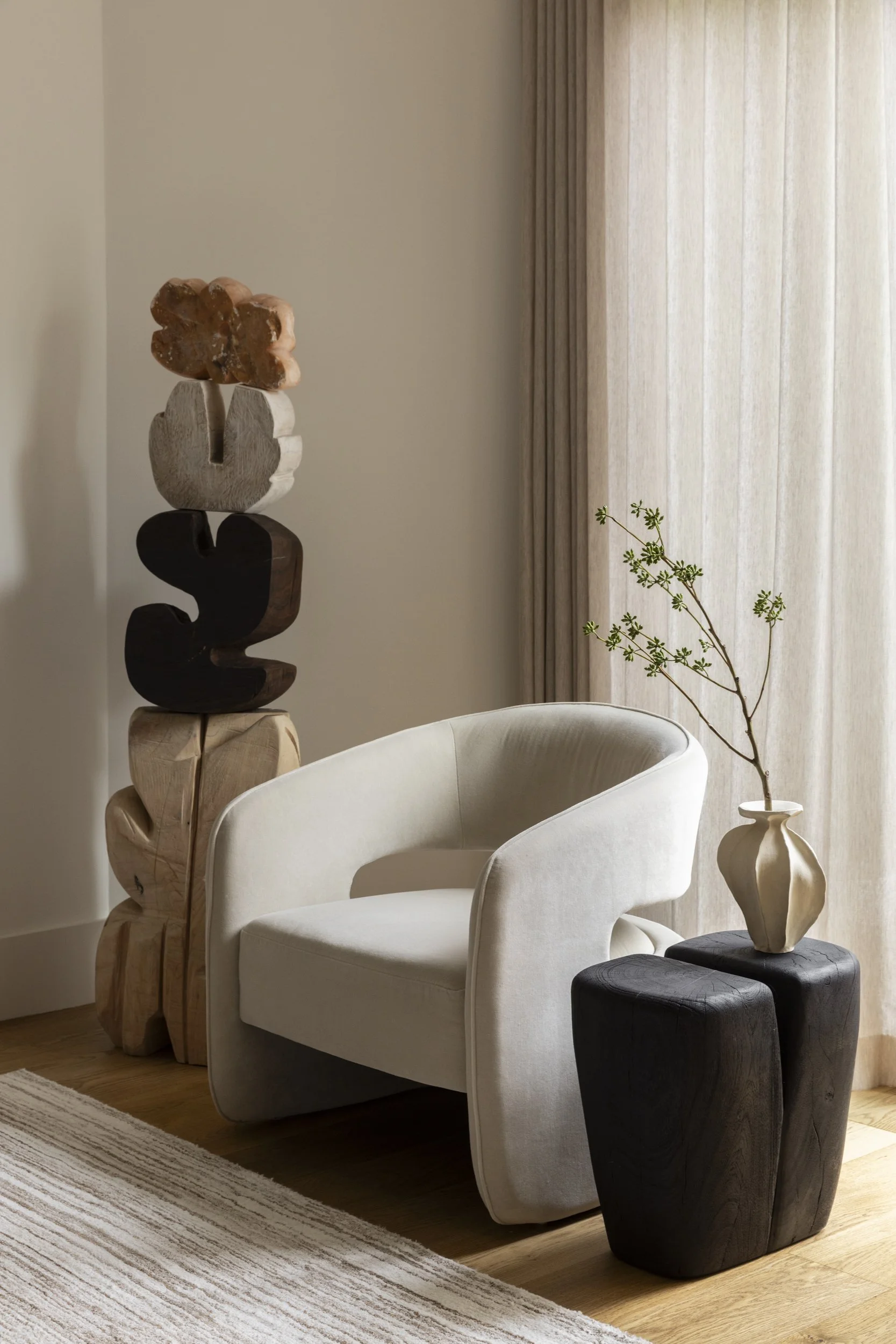 Living room corner with a modern white armchair, a black side table with a beige vase and a plant, a tall wooden sculpture with a stone resembling a flower on top, beige curtains, and a striped rug on hardwood floor.