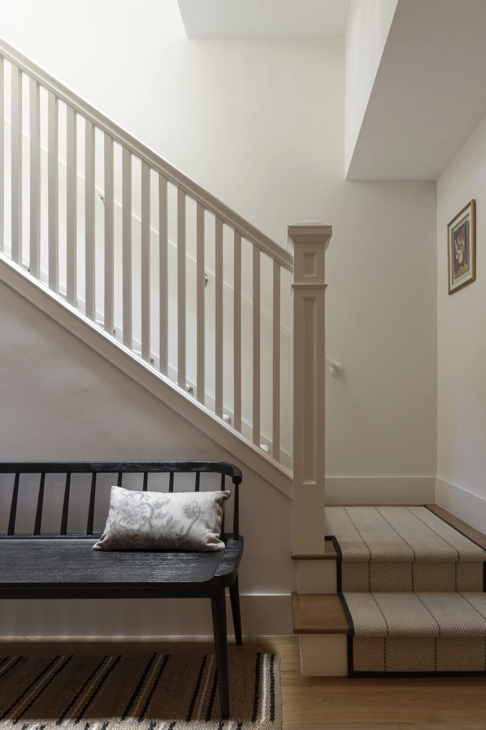 Interior view of stairs with white banister, framed artwork on wall, black bench with pillow, carpeted stairs with beige runner, and hardwood flooring.