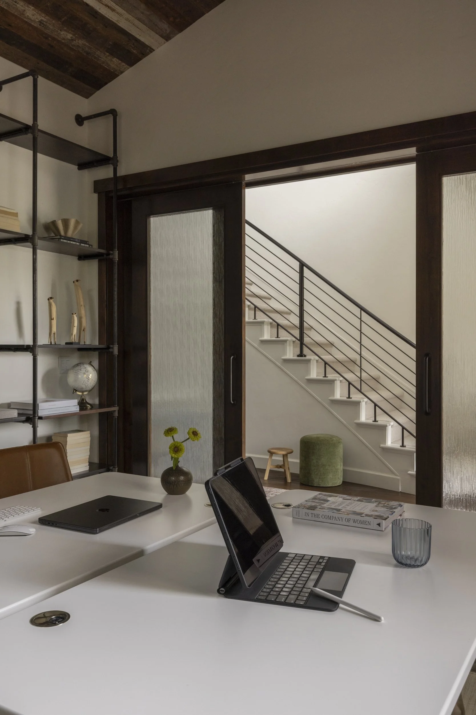 Interior of a home office with a white desk, two open laptops, a closed laptop, a tablet, a mouse, a glass of water, a book titled "In the Company of Women," a vase with yellow flowers, and a decorative glass bowl. In the background, there is a stair
