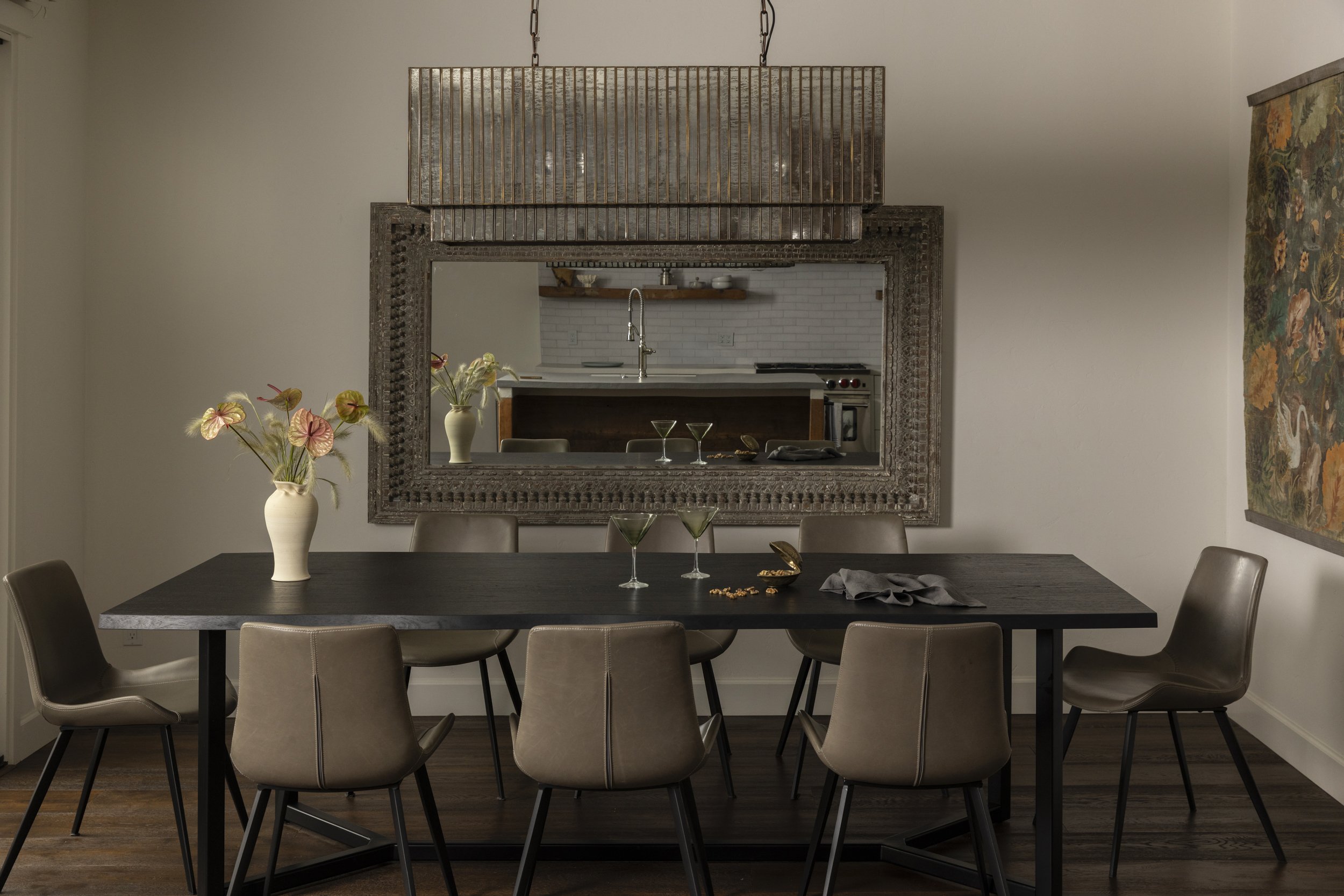 A modern dining room with a black table, ten beige chairs, a large decorative mirror on the wall, a beige vase with pink and green anthurium flowers, and a painting on the side wall. A view of a kitchen is seen through the mirror with white cabinets,