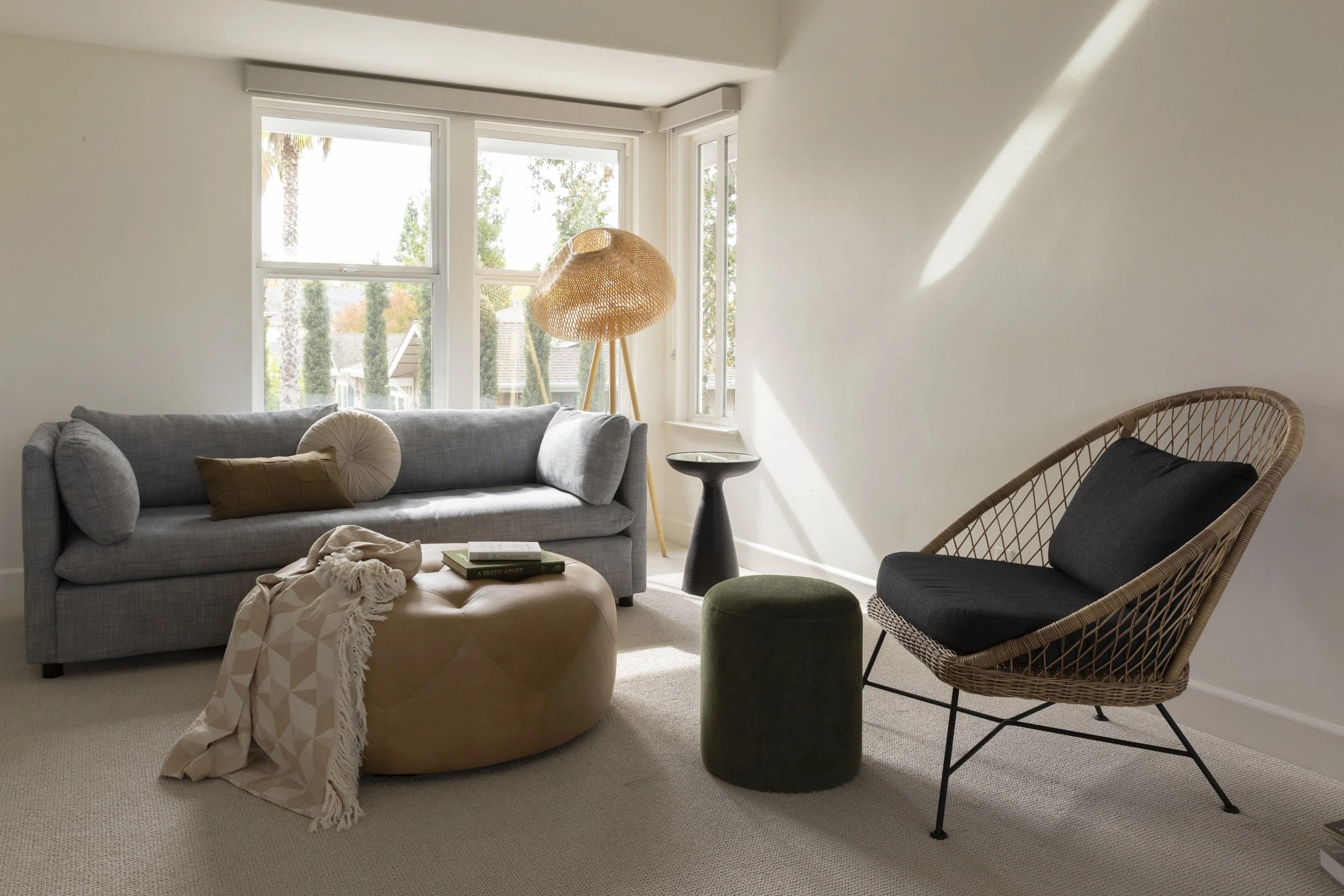 A living room with a gray sofa with cushions, a beige ottoman with a blanket, a black side table, a wicker lounge chair with a black cushion, a green ottoman, a floor lamp with a woven lamp shade, and large windows showing trees and rooftops outside.
