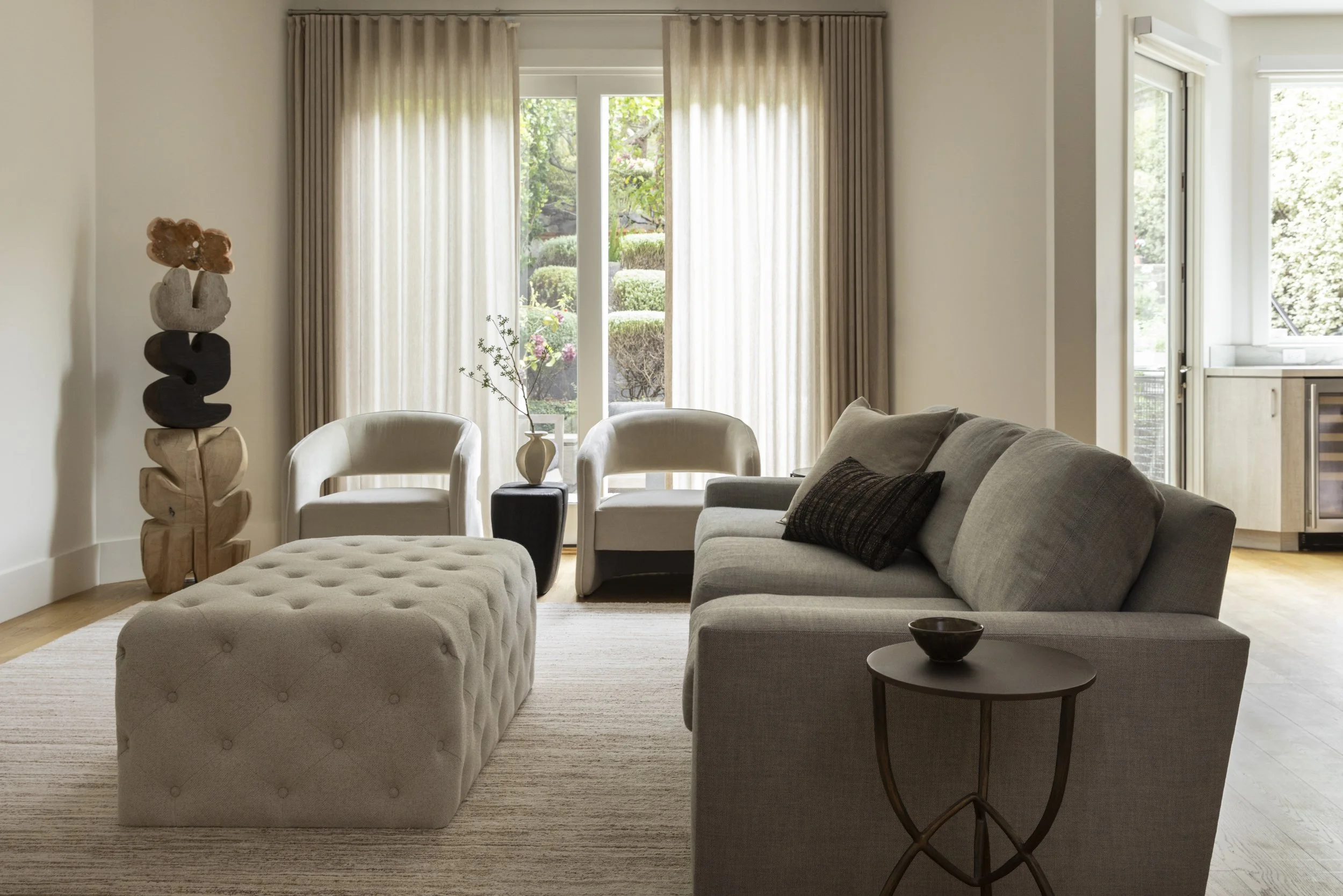 A living room featuring a beige tufted ottoman, a dark side table with a black bowl, a gray sofa with black and gray pillows, two white armchairs, a vase with pink flowers, and large windows with beige curtains showing a garden outside.
