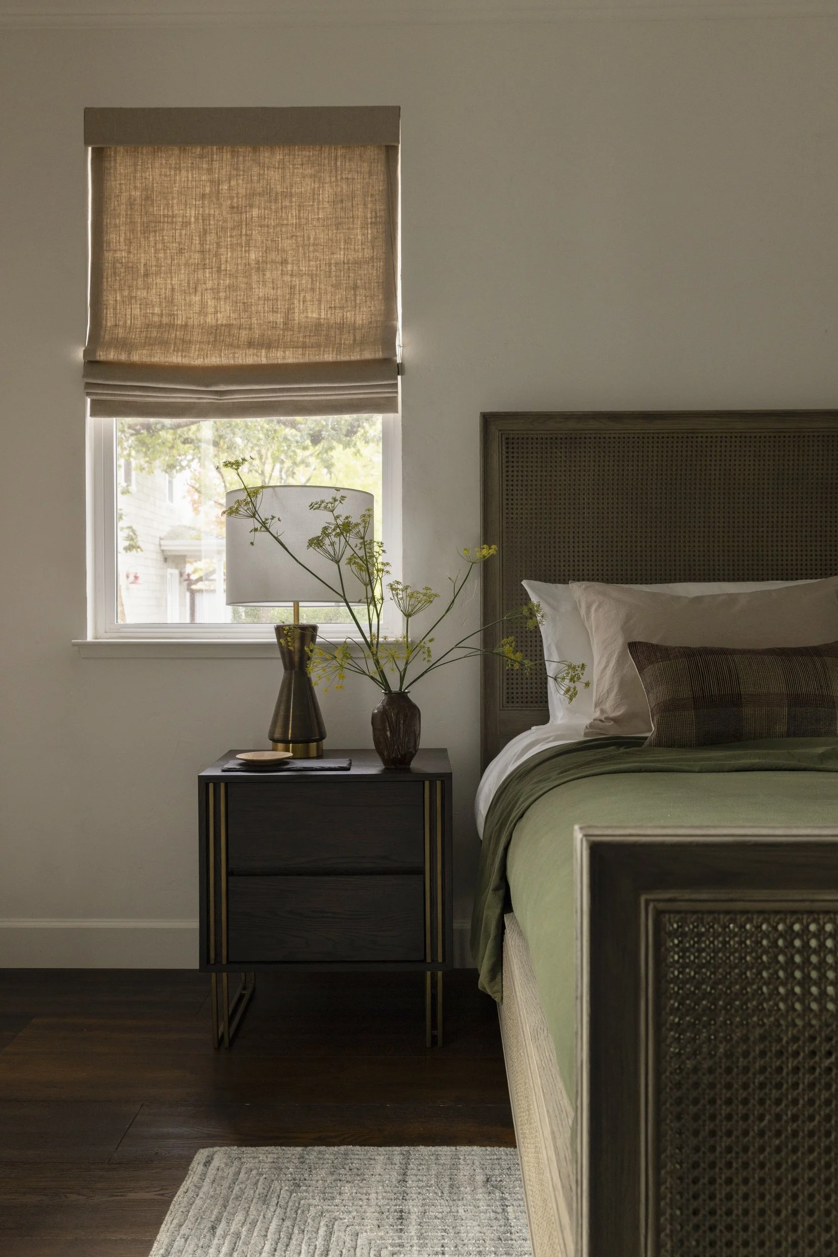A bedroom with a window covered by a beige roller shade, a black bedside table with a lamp and vase with flowers, and a bed with a green bedspread and pillows, with dark wood flooring and a gray and white rug.