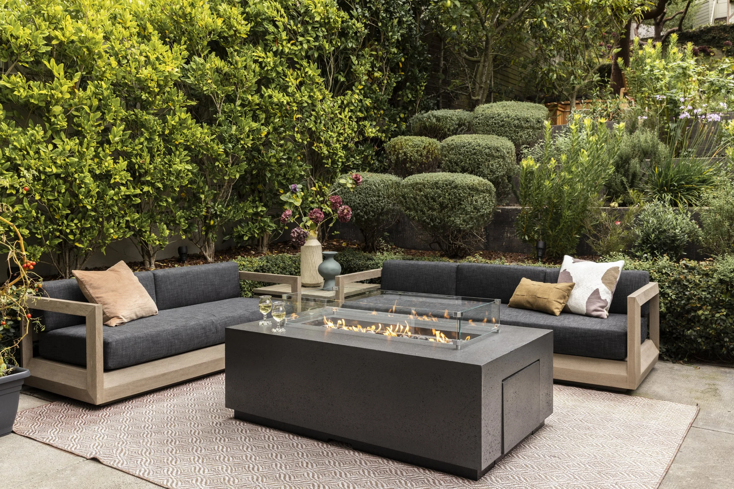 Outdoor patio with modern sectional sofa with cushions, glass-top fire pit, potted plants, lush green bushes, and trees in the background.