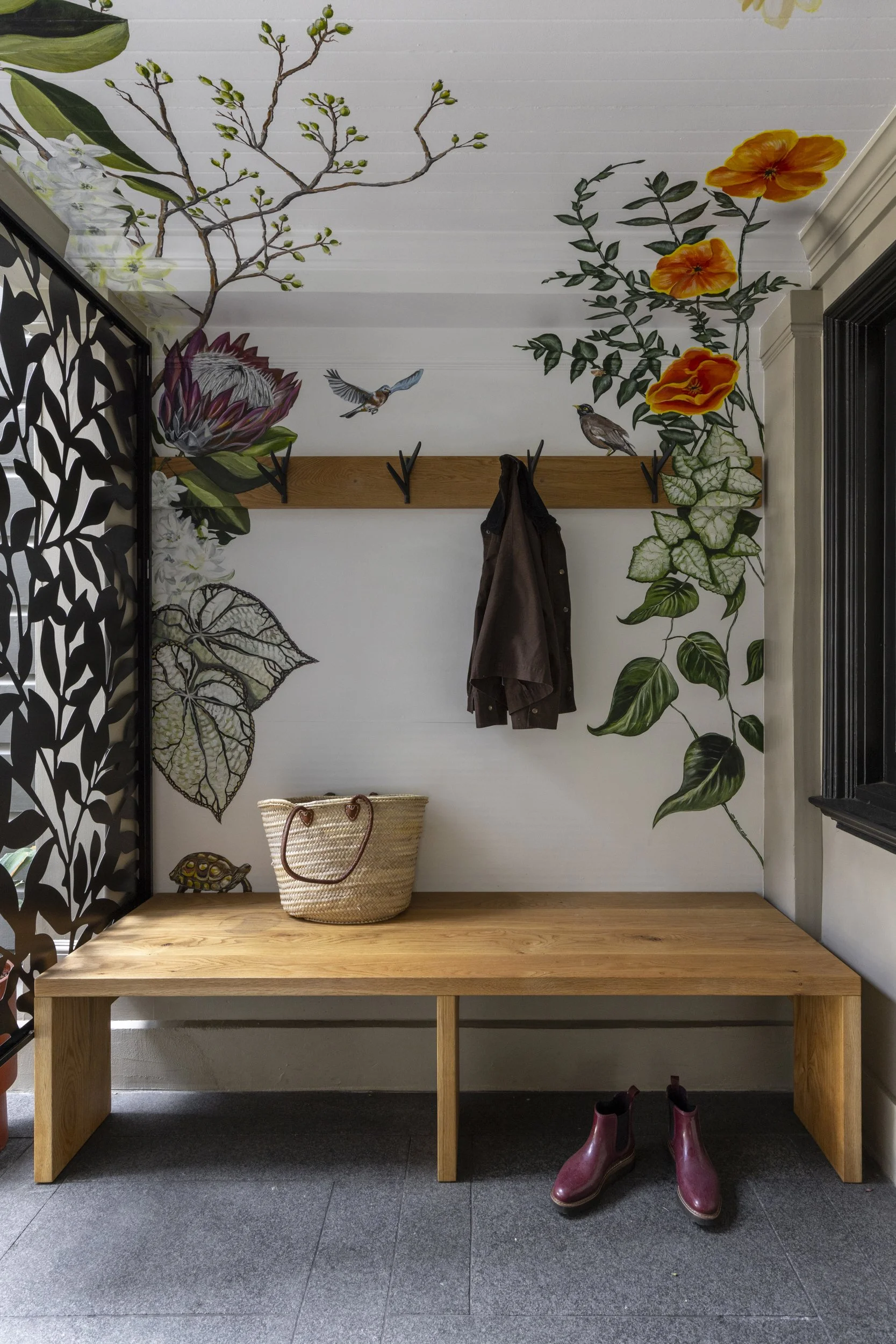 Wall mural with colorful flowers, leaves, and birds. Wooden table with a basket, a coat hanging, and pink rain boots on a dark tiled floor. Black decorative panel on the left side.