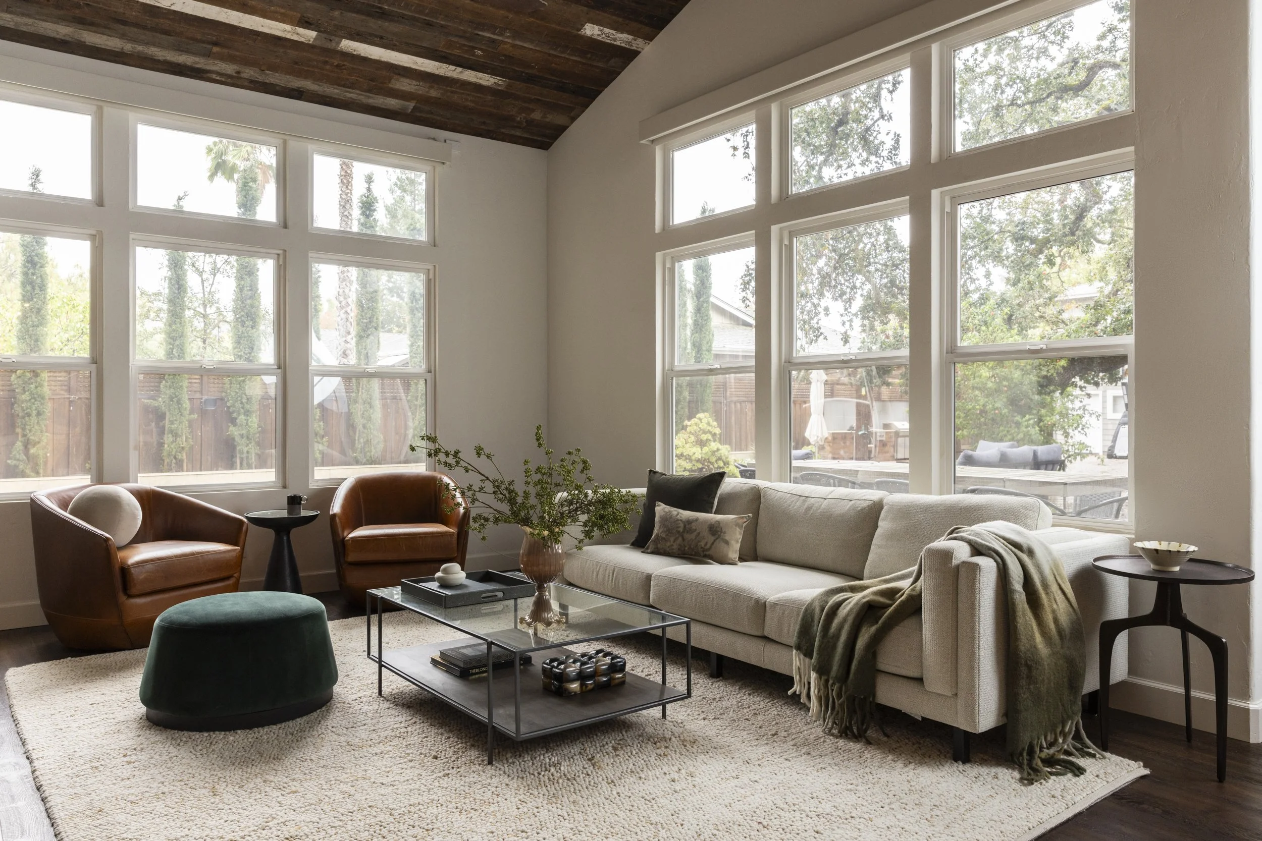 Living room with large windows, white sofa with pillows, two leather armchairs, a glass coffee table, a green upholstered ottoman, beige rug, and a potted plant, with trees visible outside.