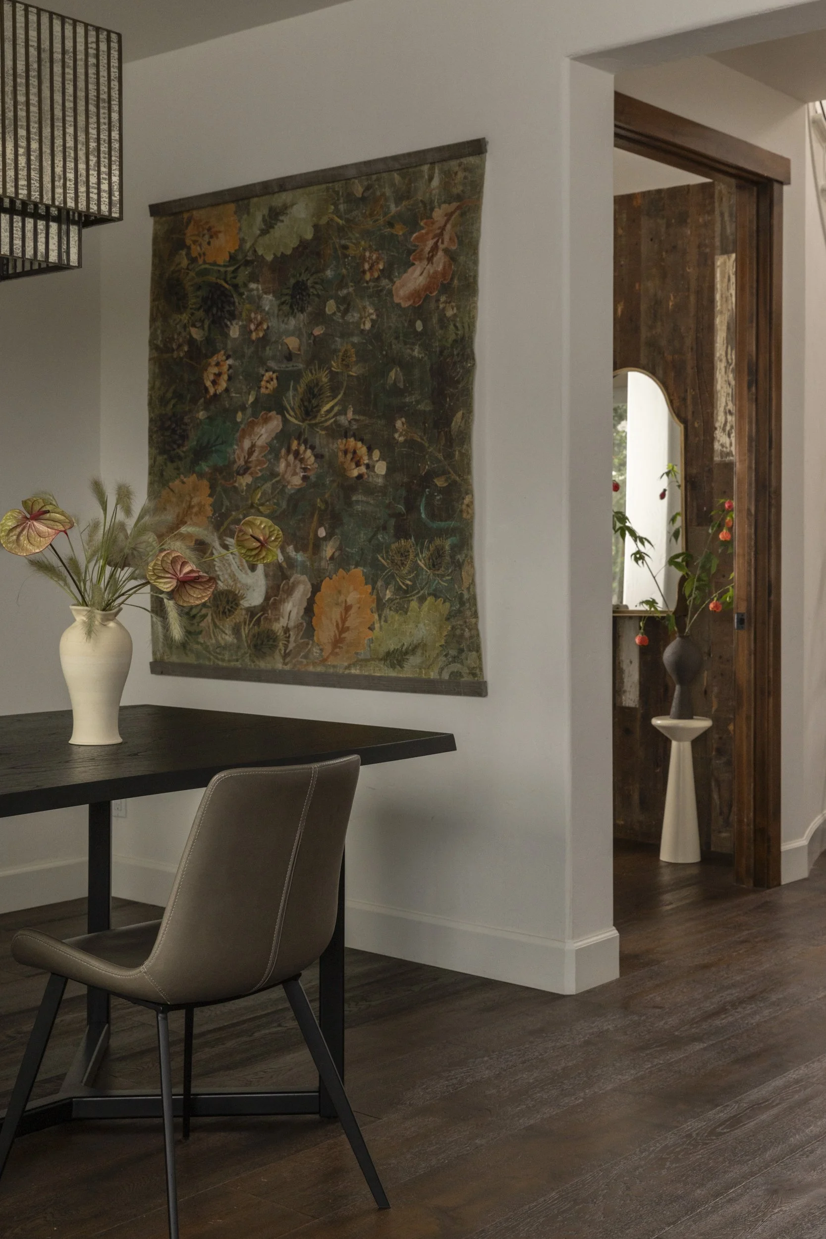 Interior view of a dining area with a dark wood table, a beige chair, a white vase with pink and green flowers, a decorative wall hanging with floral design, and a doorway with a wooden frame leading to a room with a mirror and plants.