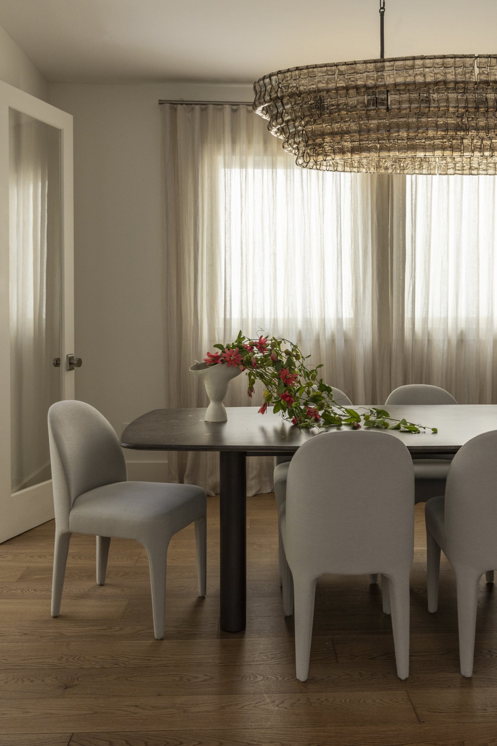 A dining room with a round dark wooden table surrounded by six light gray upholstered chairs. A white vase with pink flowers and green foliage is on the table. Light beige curtains cover the windows, letting in diffused sunlight. A large, rustic wove