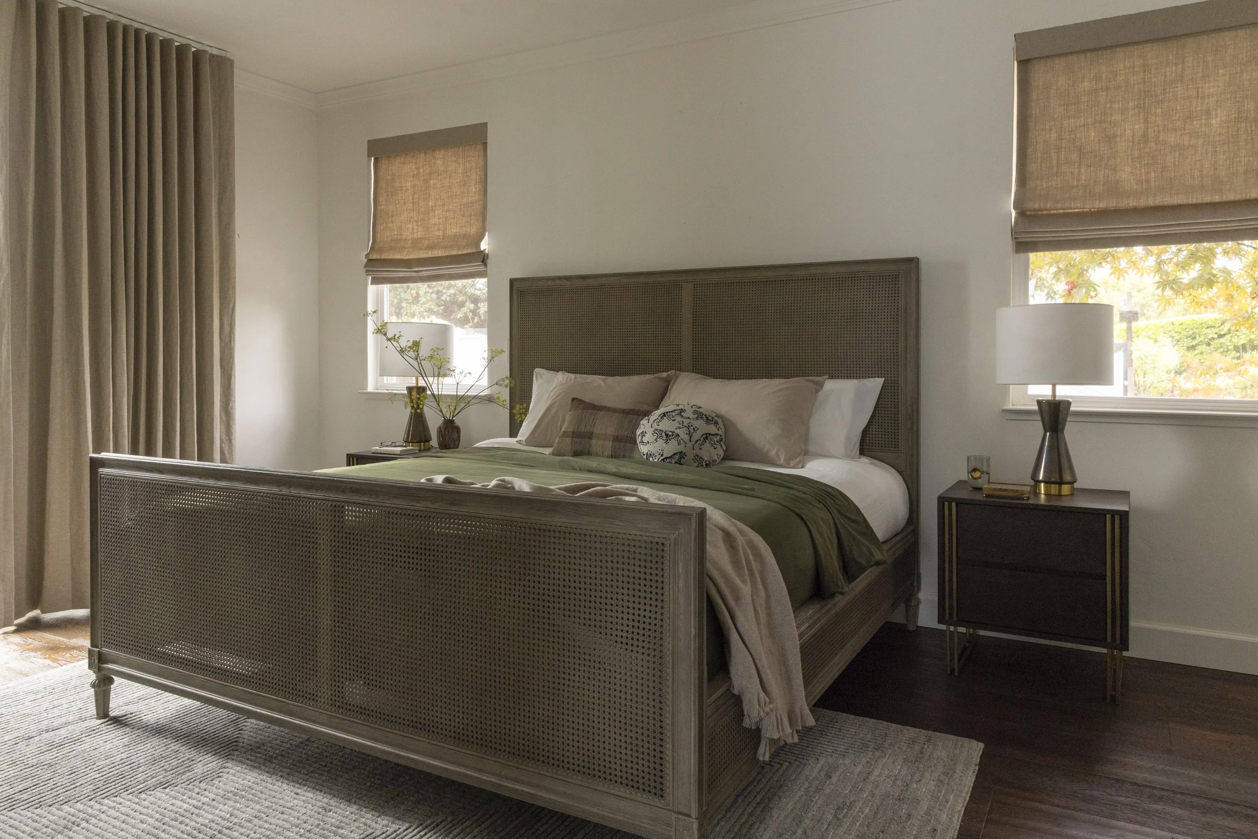 A bedroom with a wooden bed frame, beige bedding, and decorative pillows. Two windows have beige blinds, and a nightstand with a lamp and decorative items is beside the bed. There is a large area rug on the dark hardwood floor.