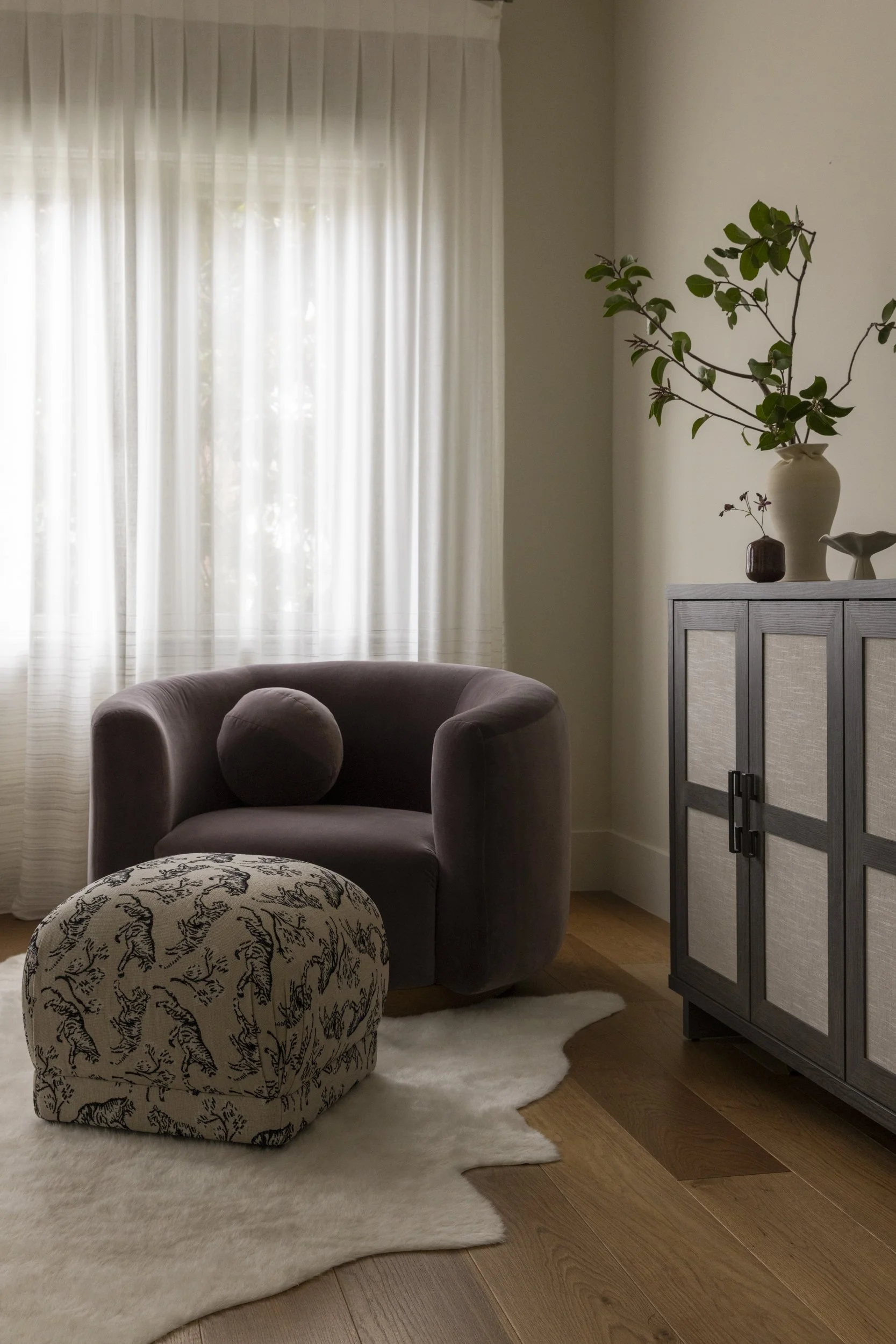 A cozy living room corner with a beige armchair, an ottoman with a black and white animal print, a white fluffy rug, a white cabinet, a large ceramic vase with green leafy branches, a smaller brown vase, and a white candle holder, with sheer white cu